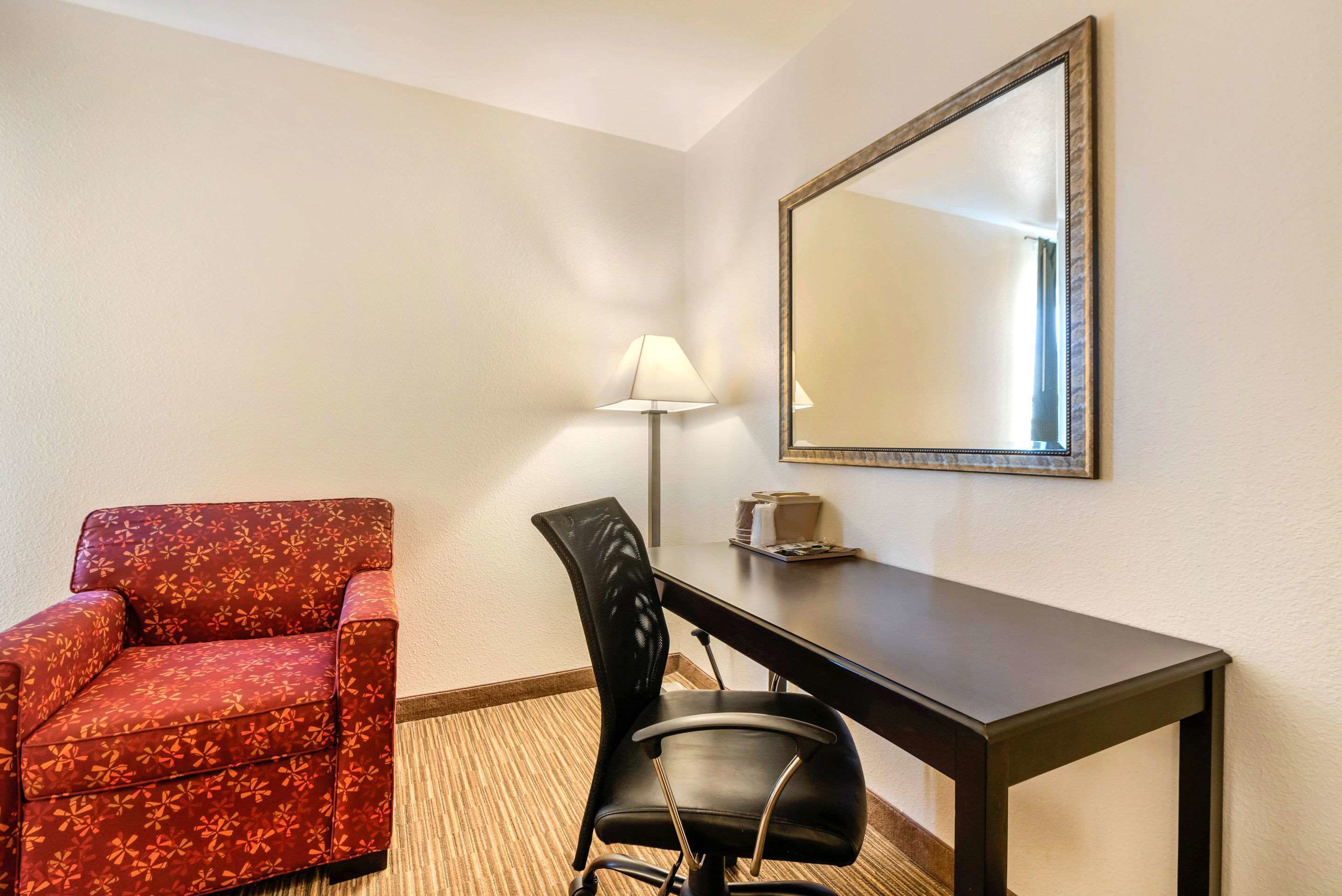 A hotel room with a red patterned armchair, a black office chair, a desk, a lamp, and a mirror on the wall.