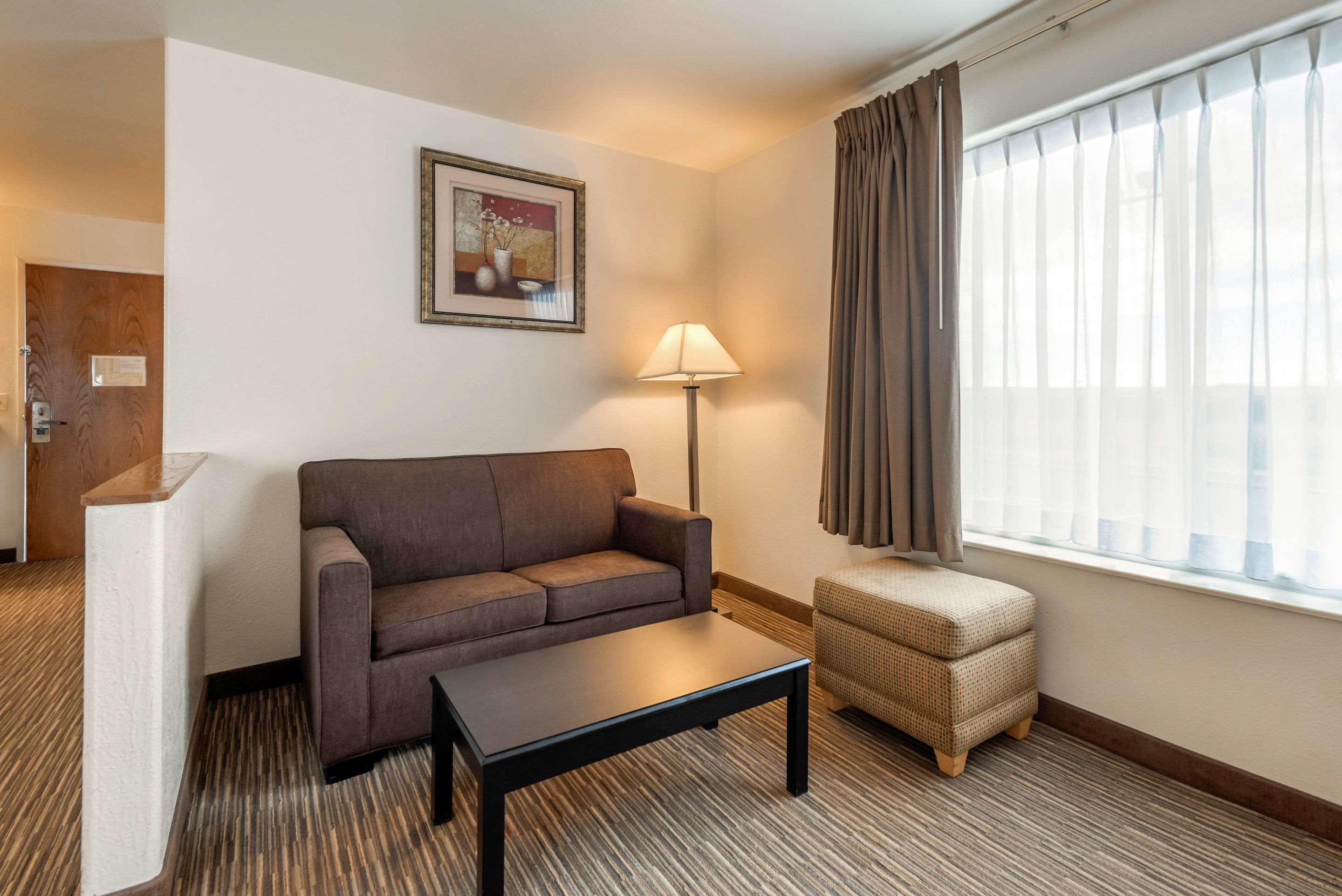 A hotel room with a sofa, a small ottoman, a lamp, a framed picture on the wall, and a window with curtains.