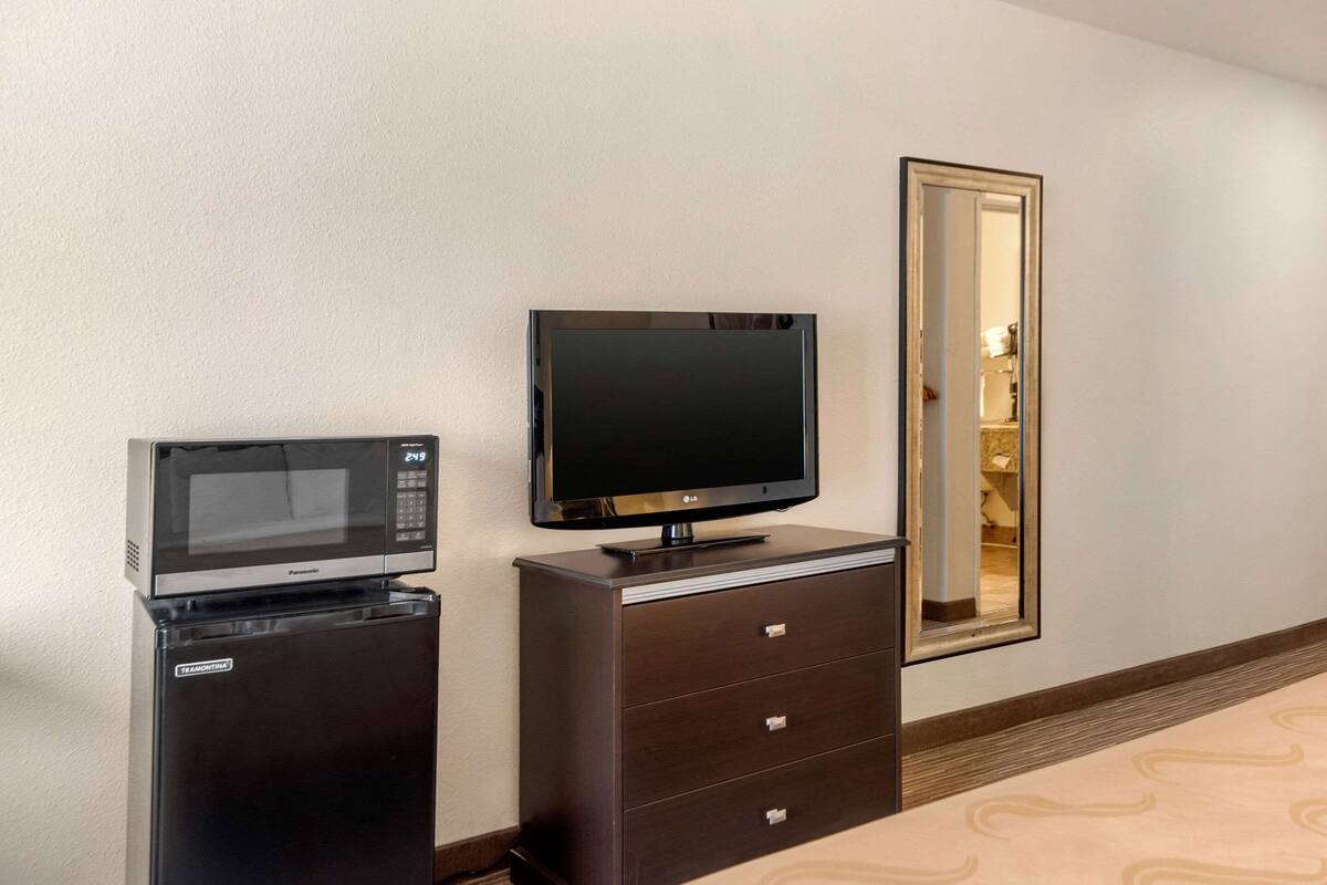A compact room with a flat-screen TV on a dresser, a microwave above it, and a mirror on the wall.