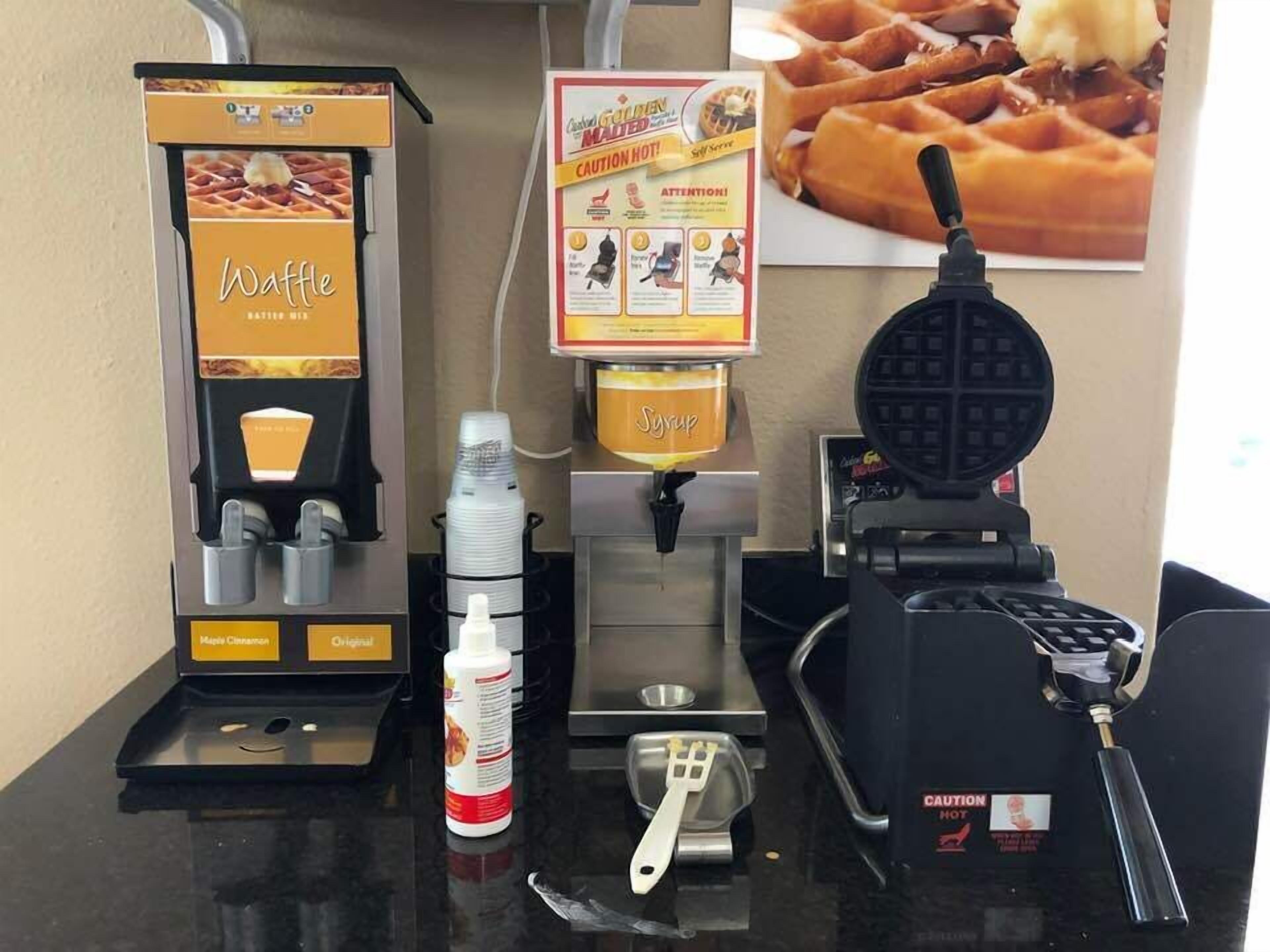 A waffle station with a syrup dispenser, a waffle maker, and a menu board.