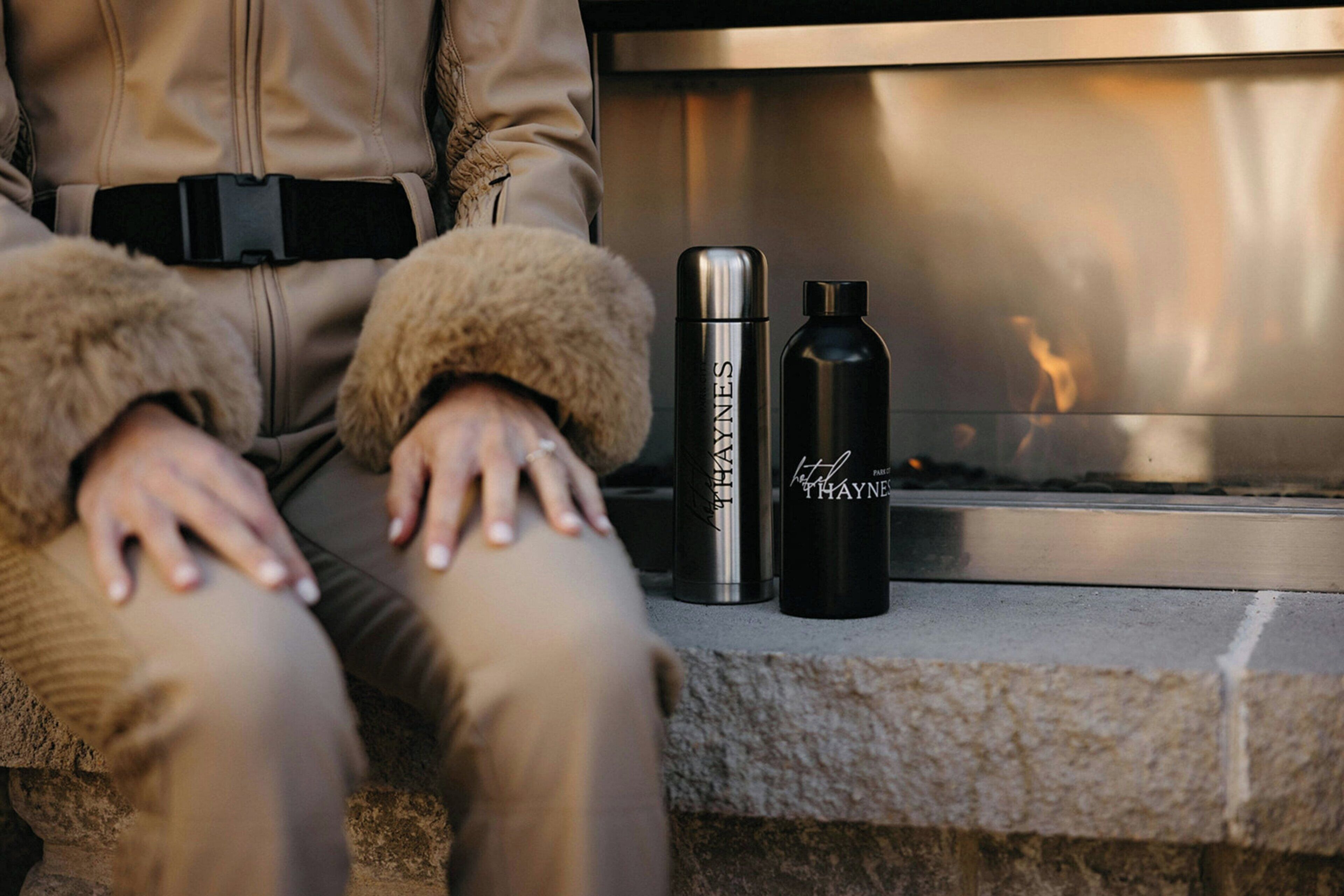 A person sitting by a fireplace with two black water bottles and a stainless steel thermos.