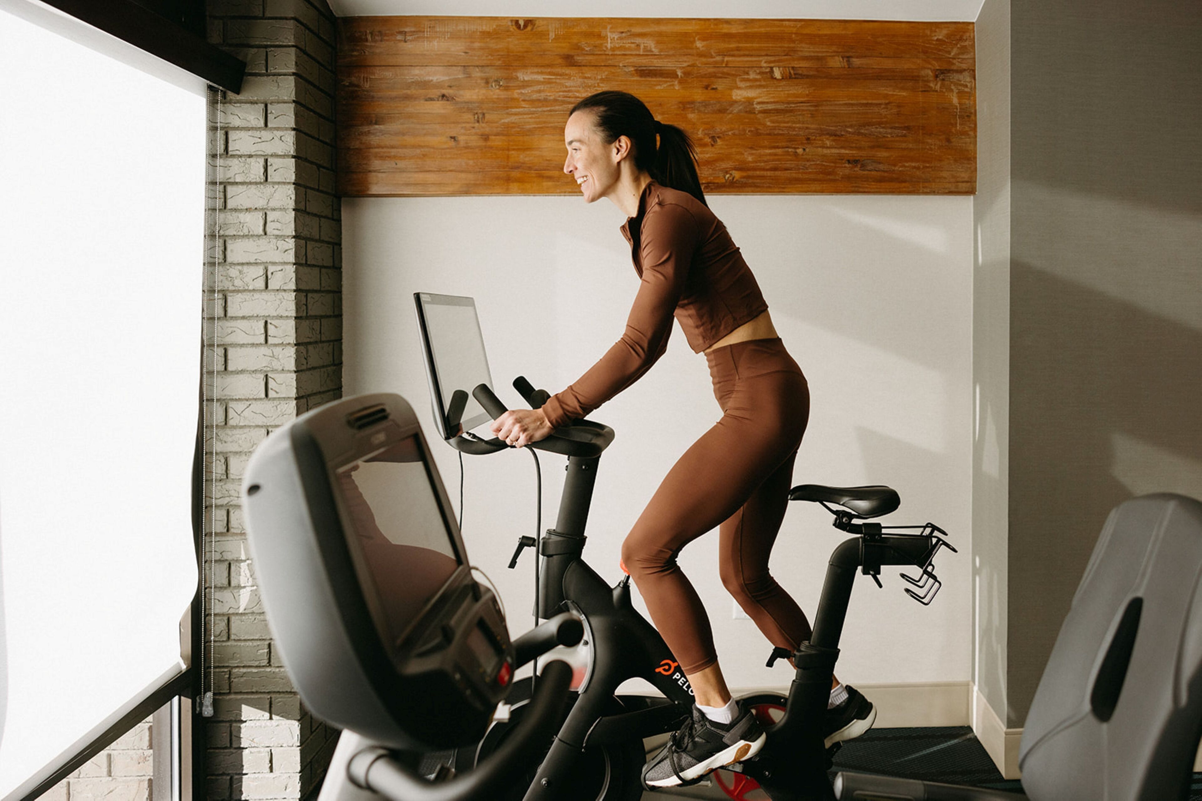 Une personne fait de l’exercice sur un vélo d’appartement dans une pièce dont un mur est en briques et un autre est recouvert de panneaux de bois.