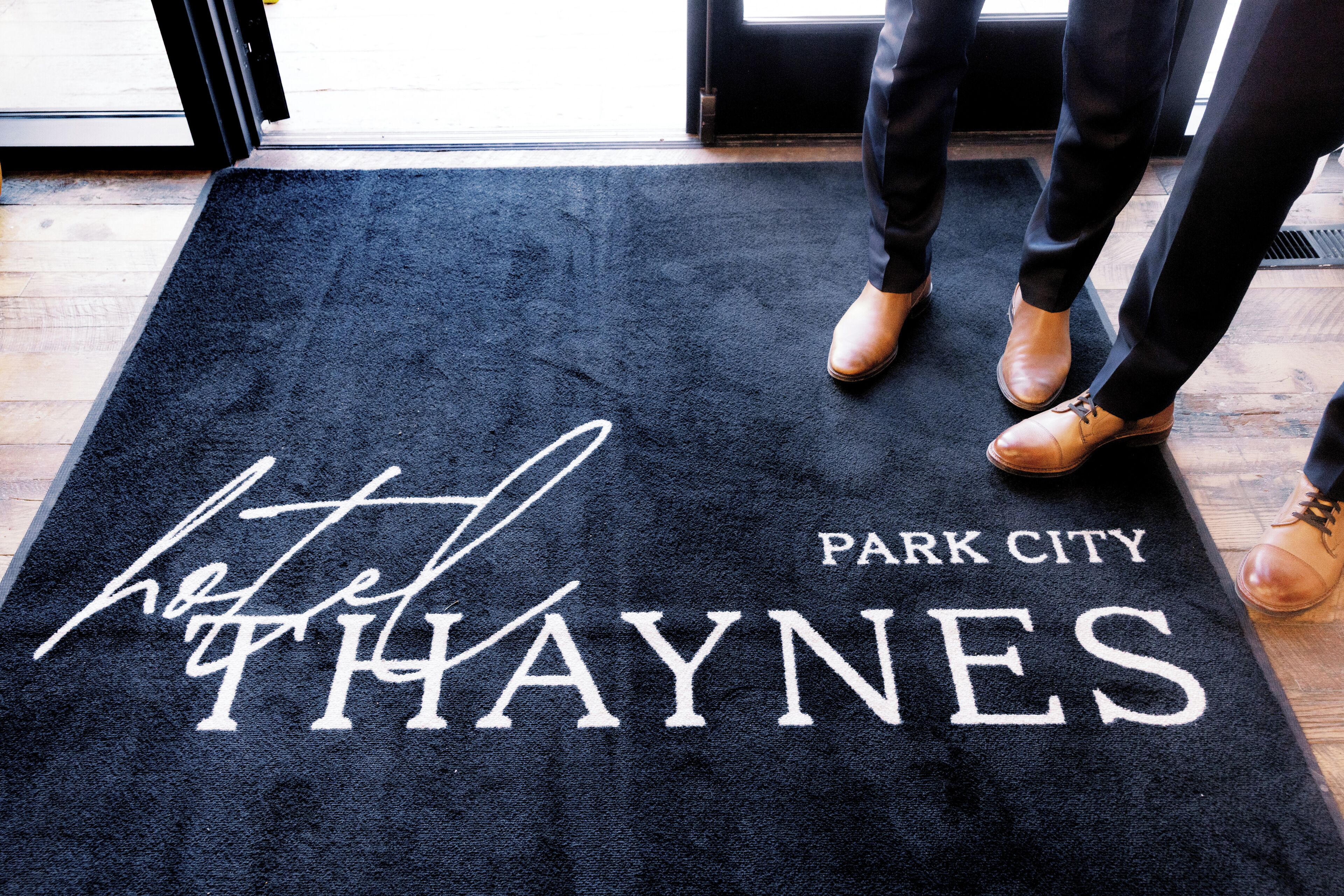 Een zwarte deurmat met de tekst 'Hotel Park City Thaynes' en een gedeeltelijk zicht op de benen van iemand met bruine schoenen.