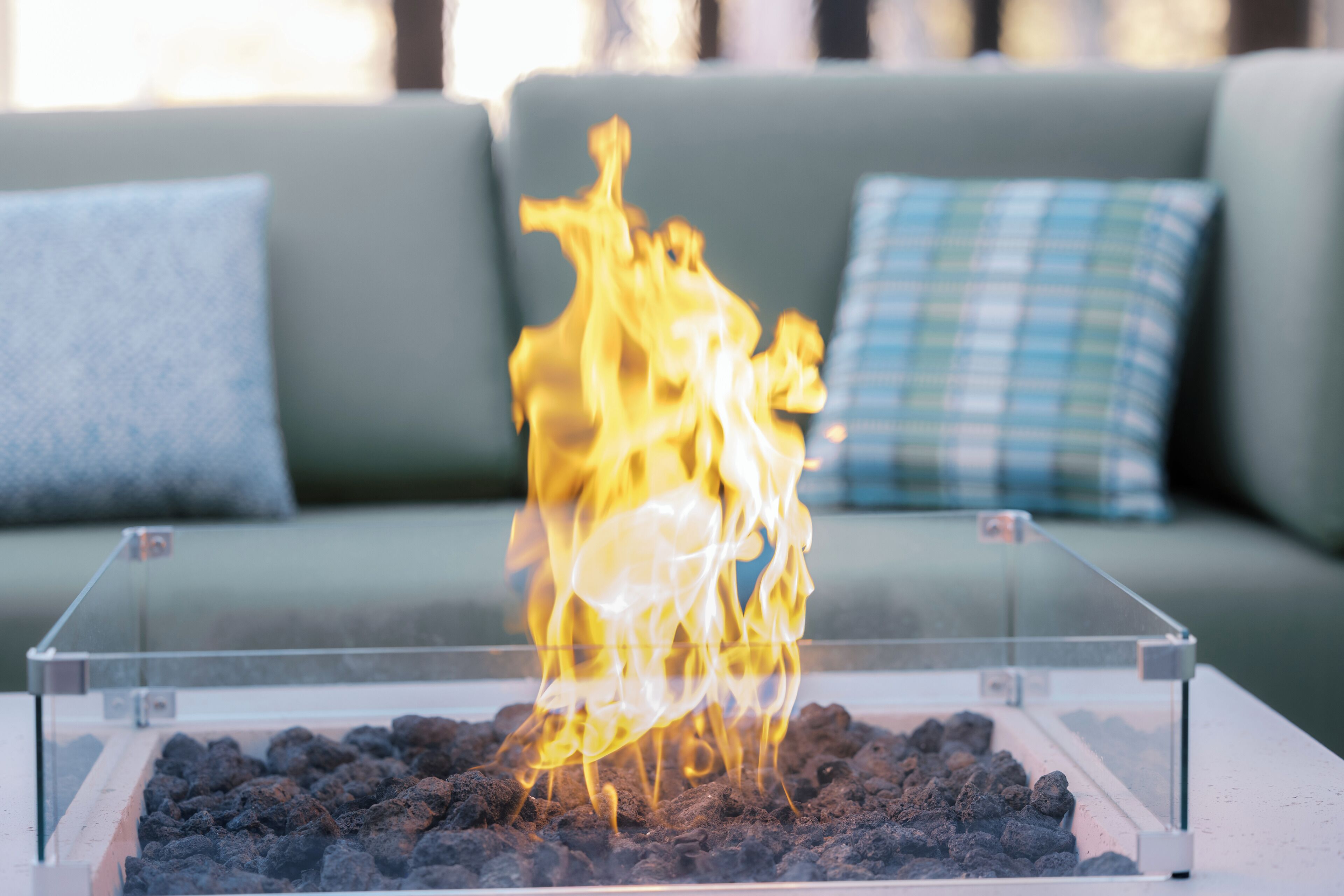 A glass fire pit with burning charcoal in the foreground, set against a green couch with a plaid cushion.