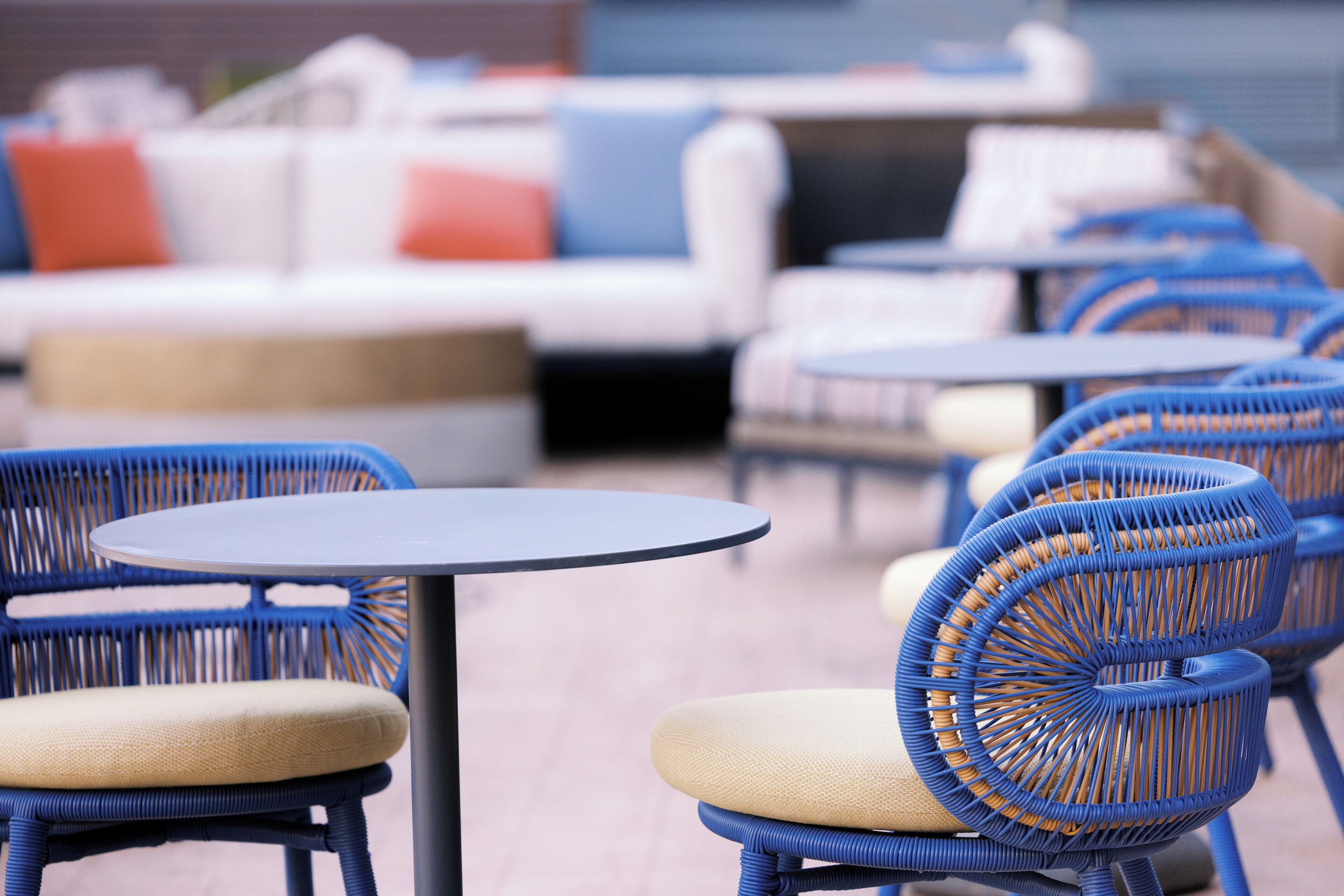 Outdoor seating with wicker chairs and round tables, a white sofa with orange and blue cushions, and a brick wall in the background.