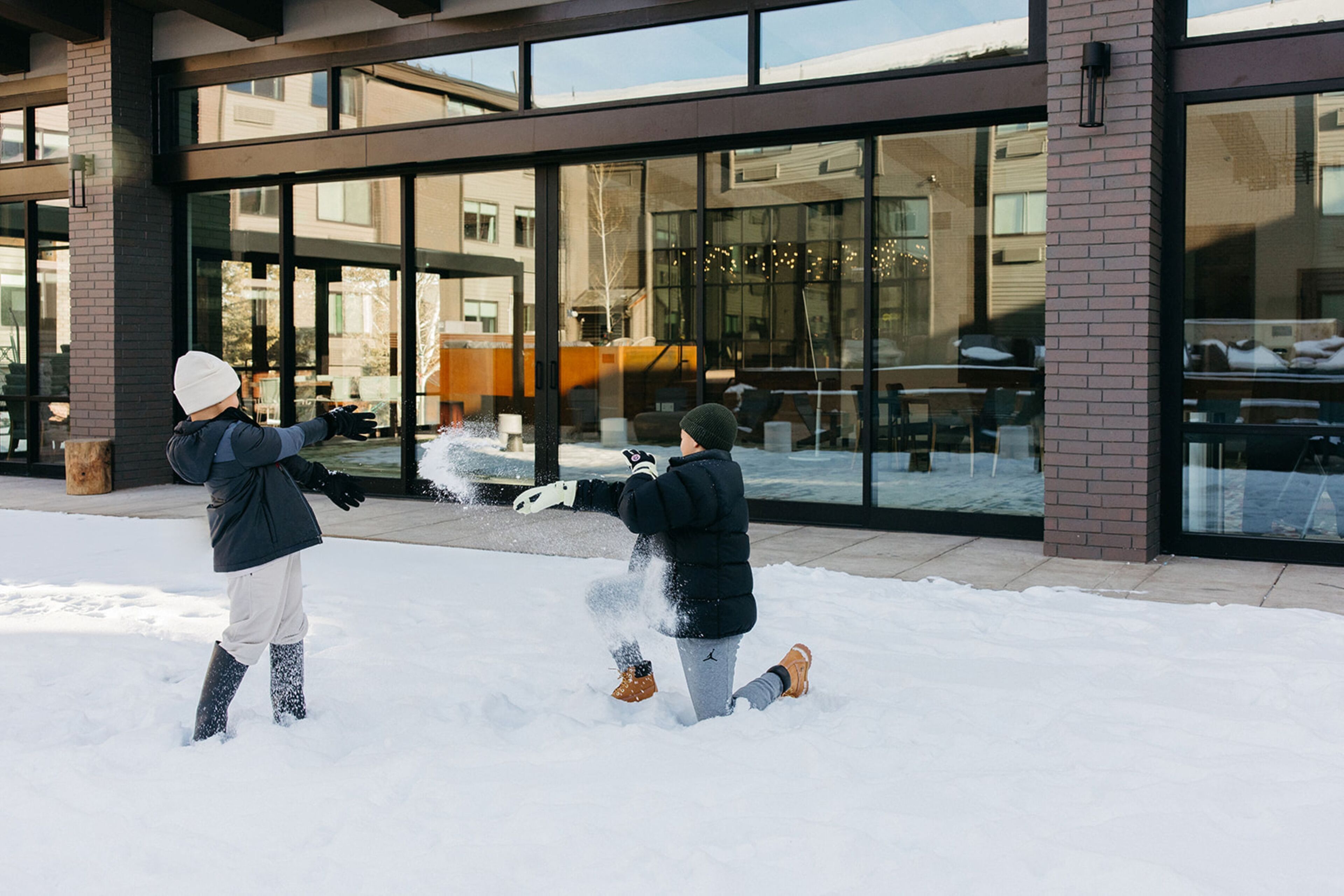 Dos personas jugando en la nieve frente a un edificio con amplios ventanales y fachada de ladrillo.