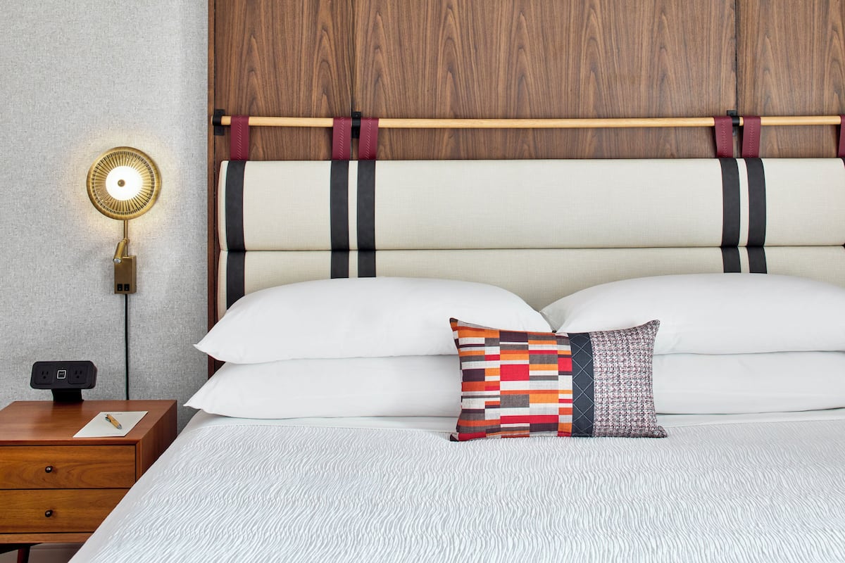 A neatly made bed with white linens and two pillows, a bedside table with a lamp, and a wooden headboard with black and red straps.