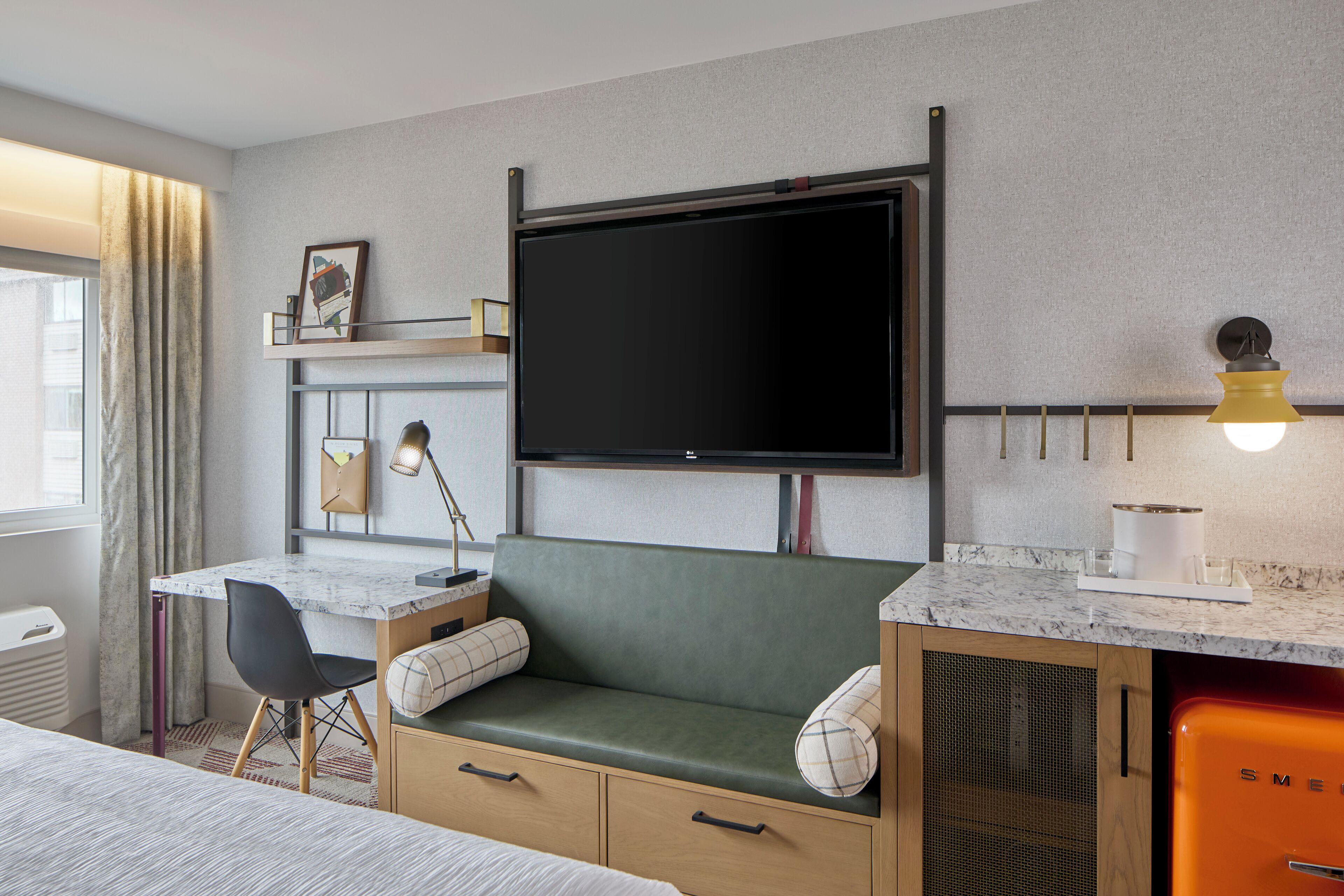 A modern hotel room with a bed, a desk with a chair, a green sofa, a television mounted on the wall, a small kitchen area with a refrigerator, and a window with curtains.