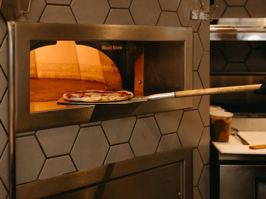 Ein Holzofen mit einer Pizza darin, ein Pizzaschieber und ein Schneidebrett aus Holz.