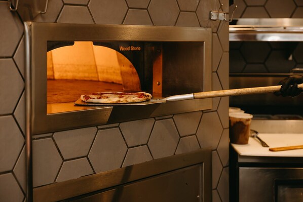 A wood-fired pizza oven with a pizza inside, a pizza peel, and a wooden cutting board.