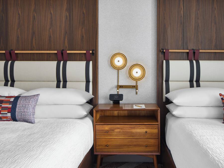 Een tweepersoonsbed met een houten hoofdbord, een nachtkastje met een lamp en een wandlamp.