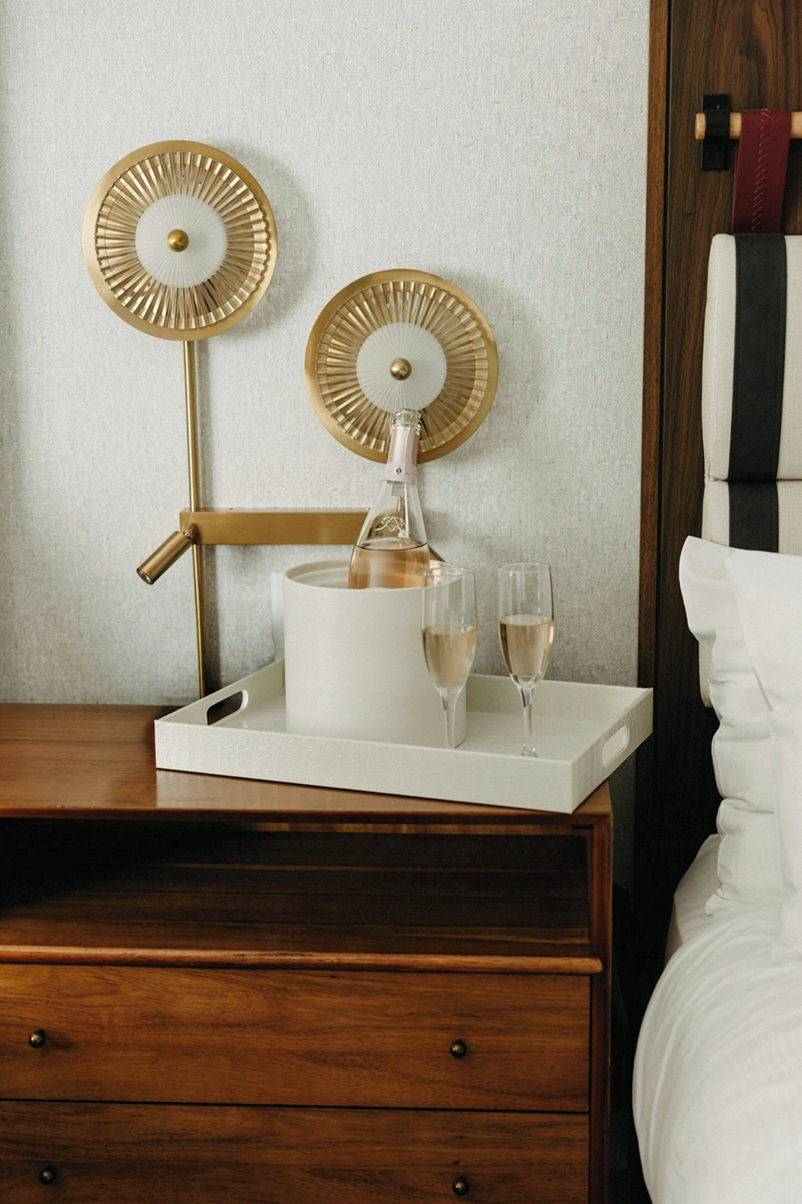 Une chambre d’hôtel avec une table de chevet en bois, un plateau blanc sur lequel se trouvent une bouteille de champagne, deux verres et deux horloges décoratives murales.