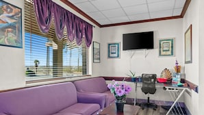 Ein Wohnzimmer mit einem violetten Sofa, einem kleinen Tisch mit Blumen, einem schwarzen Bürostuhl, einem an der Wand befestigten Fernseher und gerahmten Bildern.