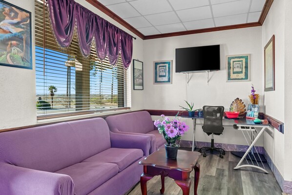 A living room with a purple sofa, a small table with a vase of flowers, a black office chair, a TV mounted on the wall, and framed pictures.