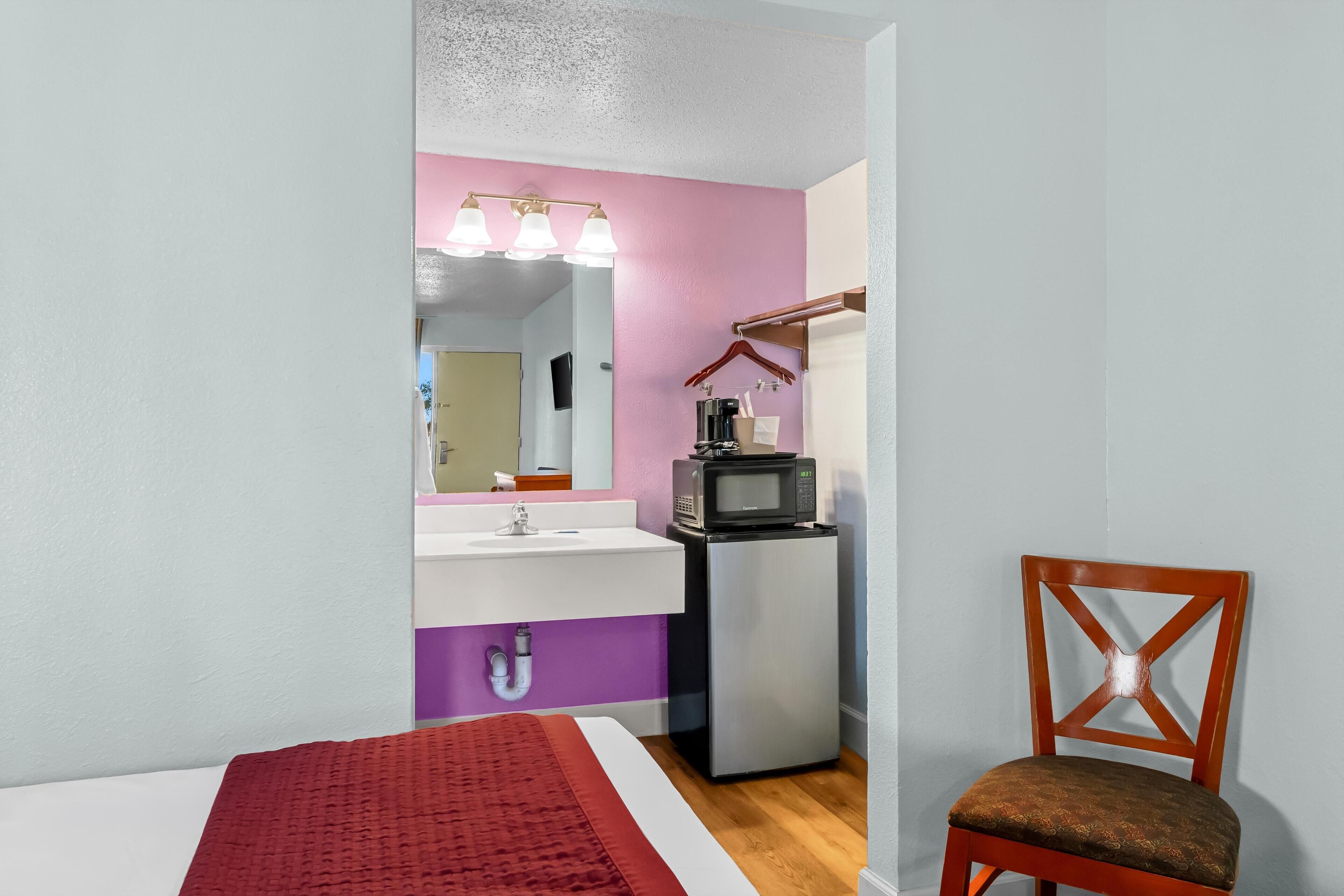 Een slaapkamer met een bed, een rode deken, een houten stoel, een kleine kitchenette met een magnetron en een wastafel.