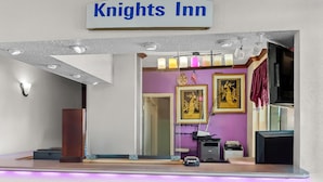 Ein Empfangsbereich mit einem Schild, auf dem 'Knights Inn' steht, einem Empfangstisch, einem Spiegel und einer violetten Wand mit gerahmten Kunstwerken.