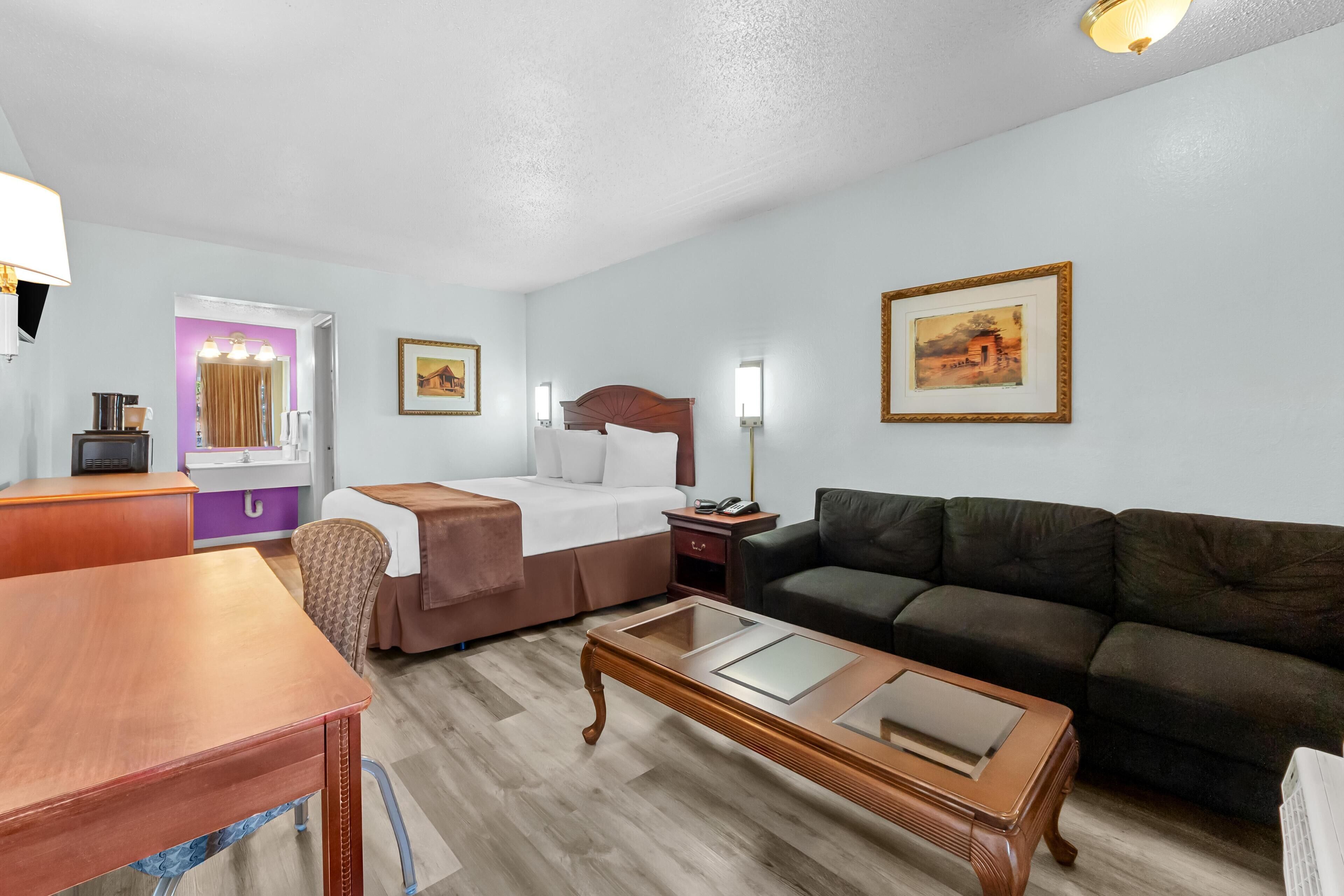 Habitación de hotel con cama, sofá, mesa de madera, escritorio y baño con tocador iluminado en morado.