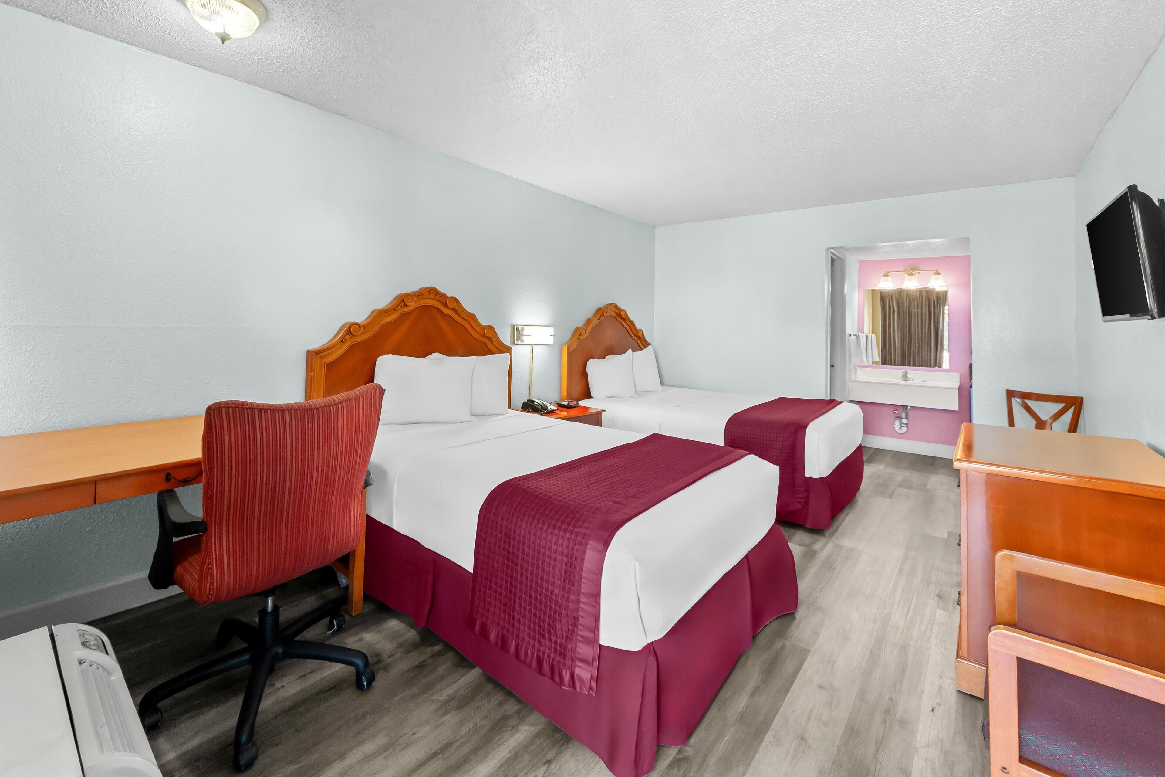 A hotel room with two beds, a desk, a chair, a TV, and a bathroom with a mirror and lights.