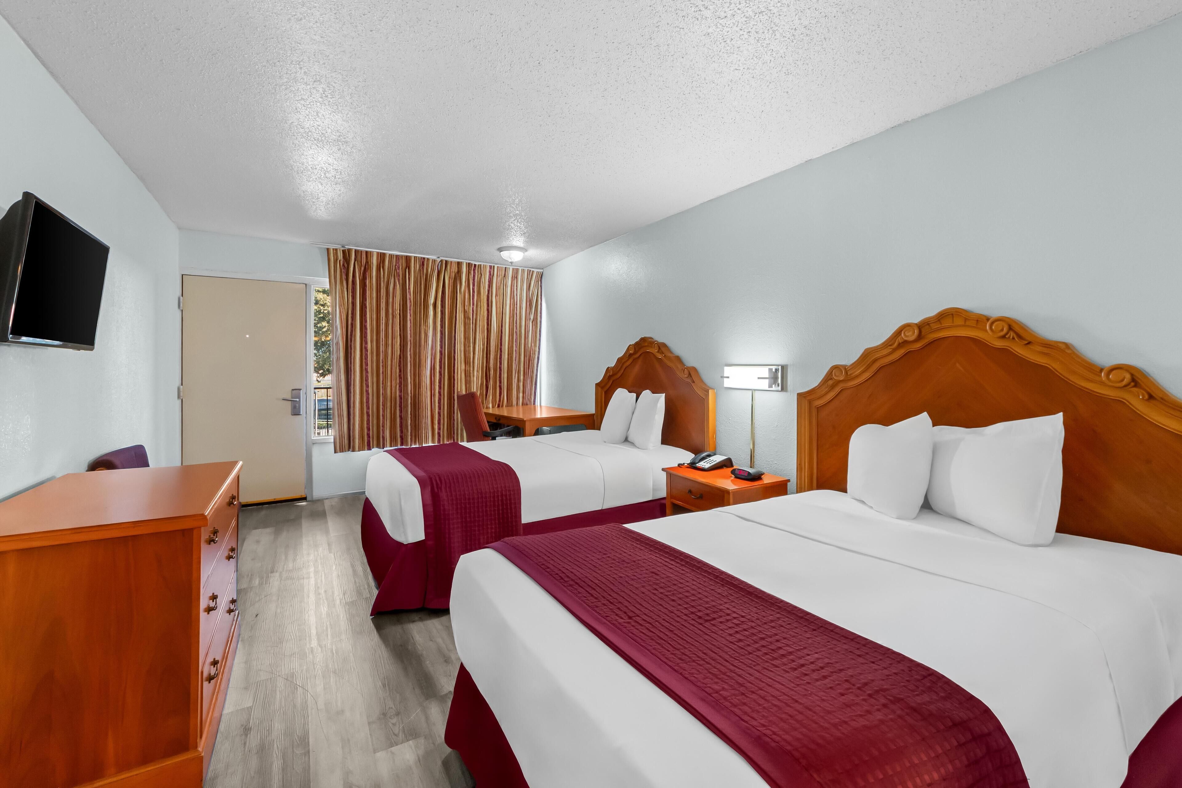 A hotel room with two beds, a wooden desk, a television, and a window with curtains.