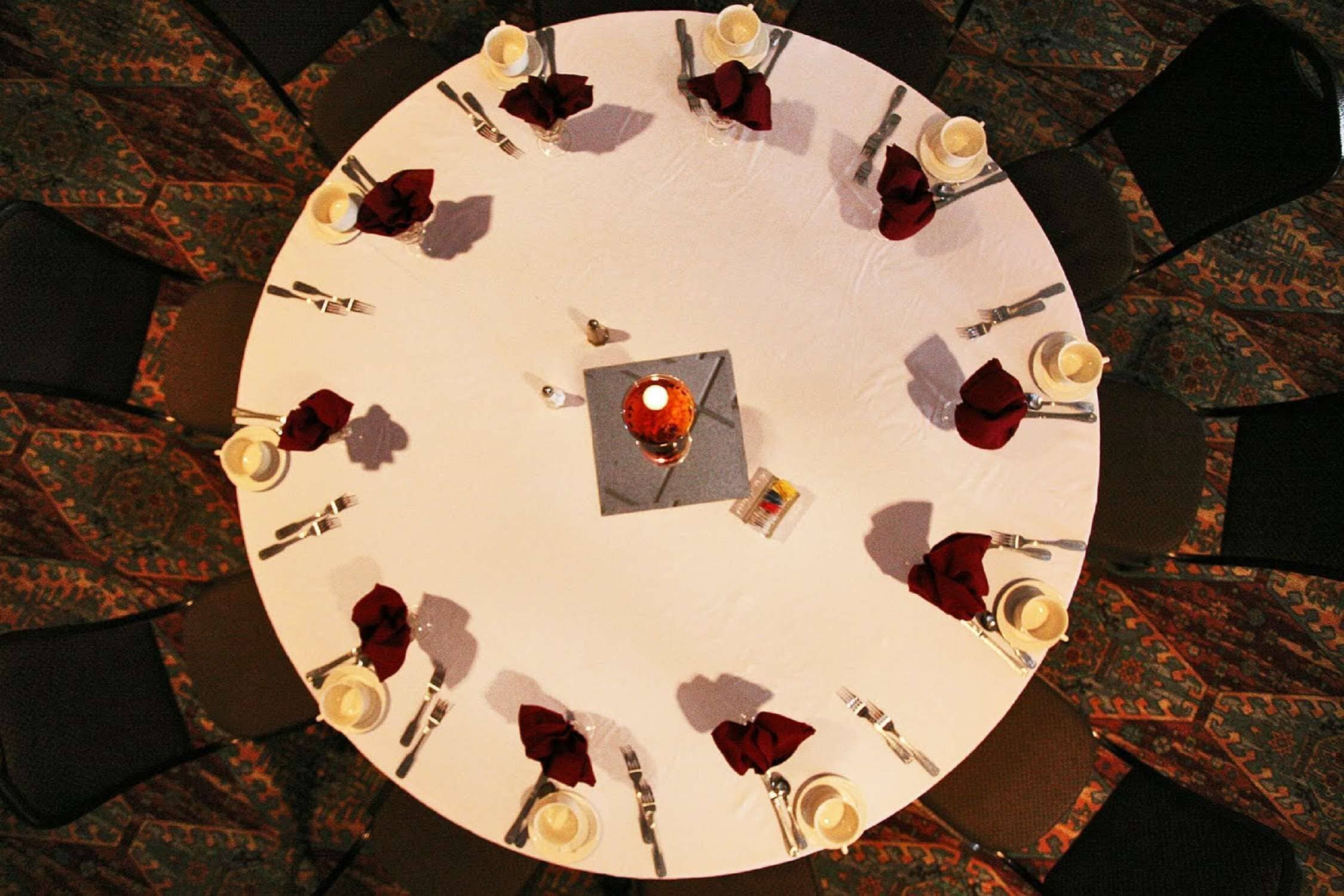 Une table ronde entourée de huit chaises, chacune recouverte d’une nappe blanche, agrémentée de serviettes rouges et de couverts en argent. Une petite table carrée, placée au centre, porte une bougie et un menu.