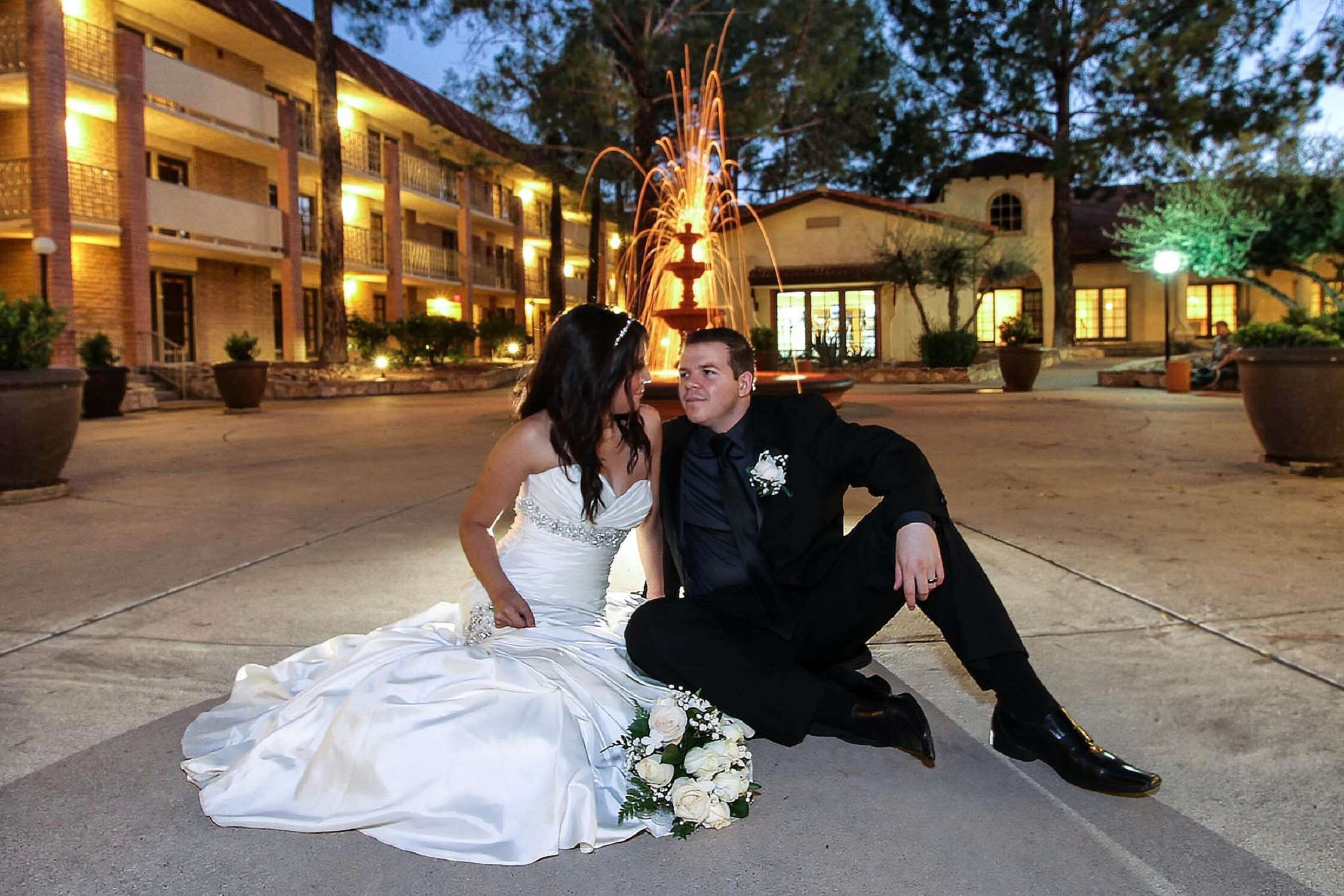 A wedding couple seated on a concrete surface in front of a building with a fountain and decorative lights.