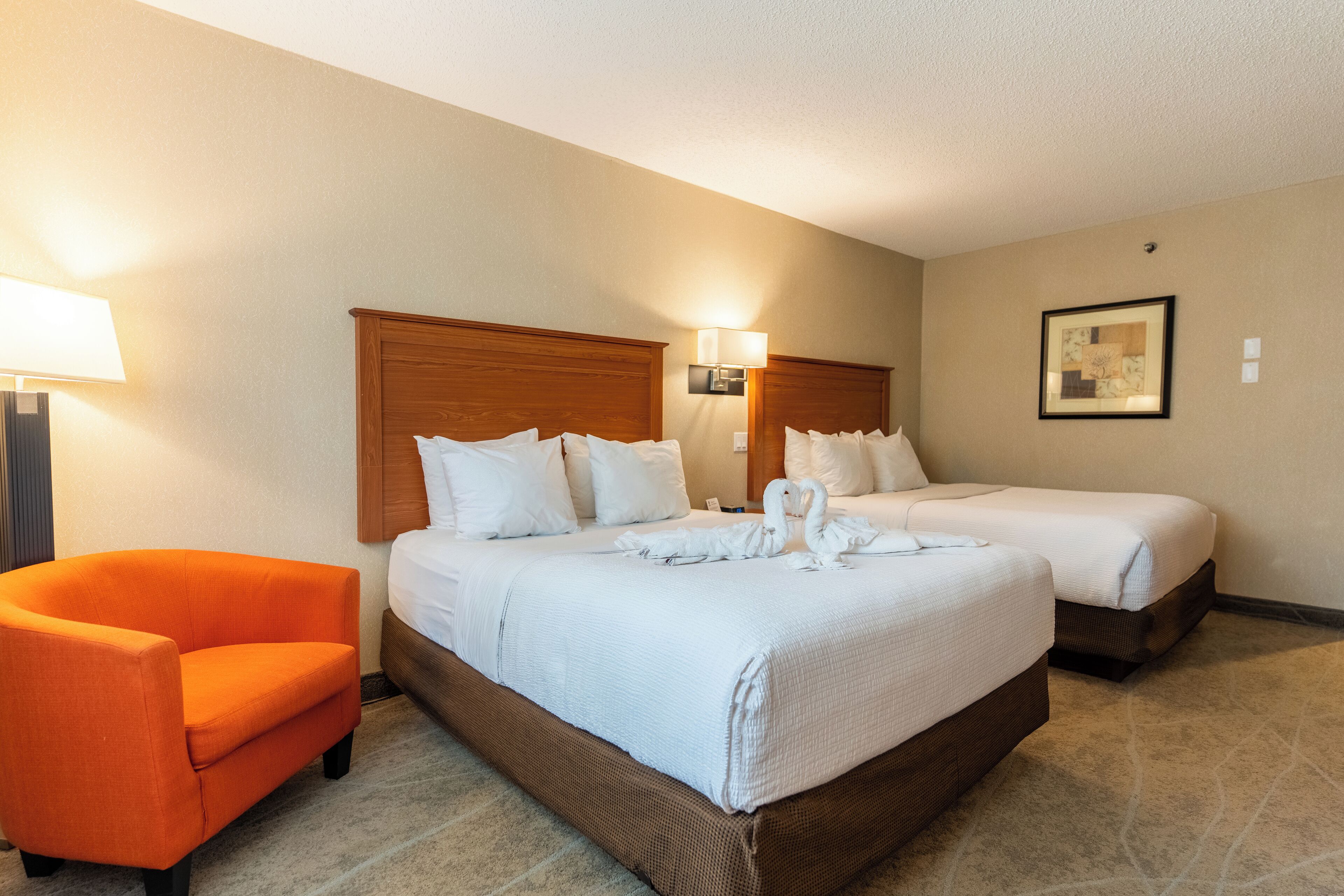 A hotel room with two beds, a wooden headboard, a white bedspread, a pair of white towels, a small orange chair, and a framed picture on the wall.