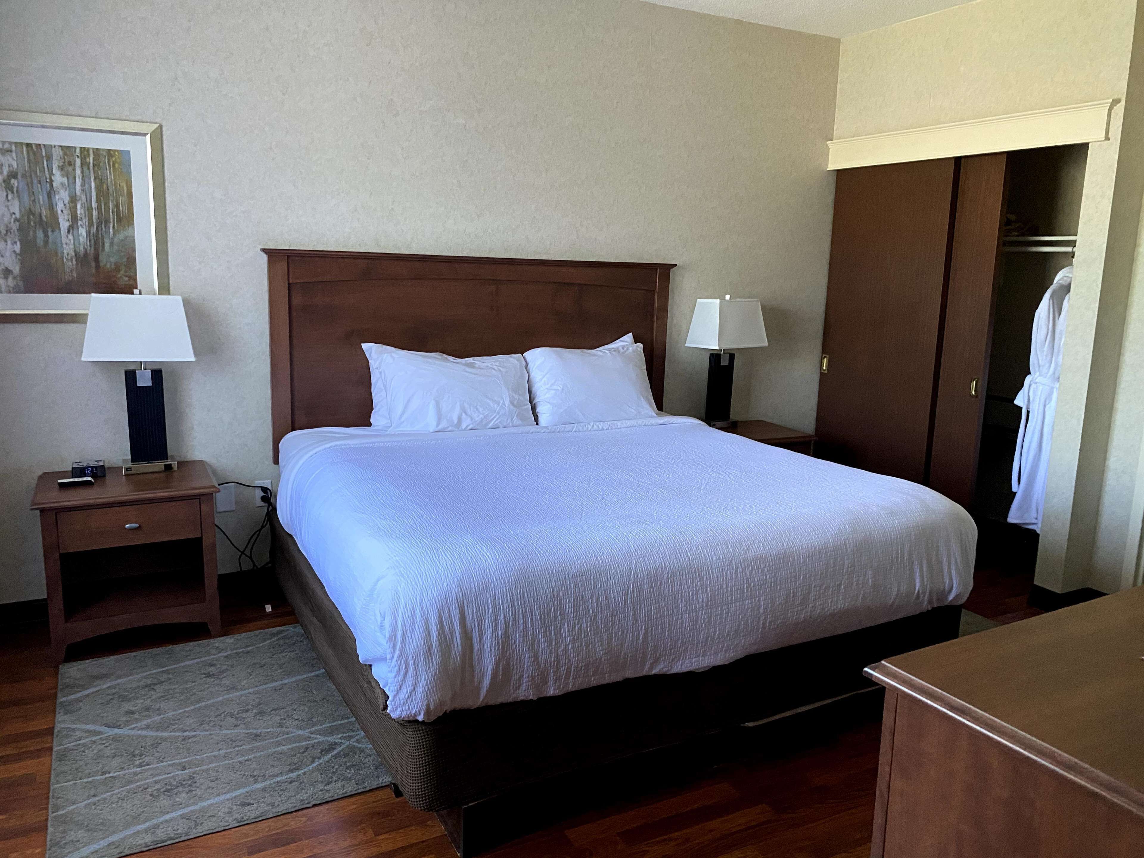 A neatly made bed in a hotel room with a wooden headboard, two bedside tables with lamps, a nightstand, a robe hanging on a door, and a painting on the wall.
