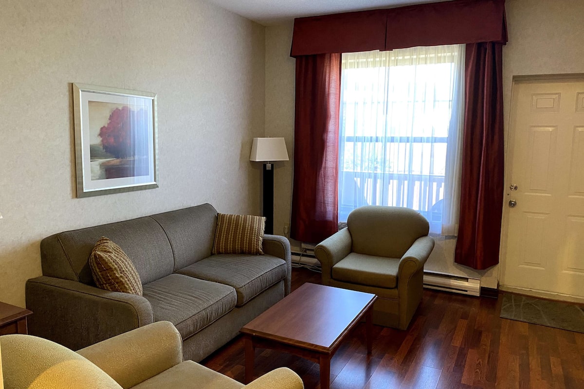 A living room with a sofa, armchair, and coffee table. There is a lamp, a framed picture on the wall, and a door.