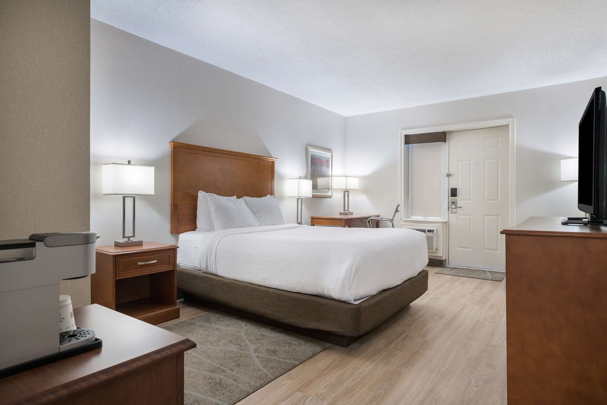 A hotel room with a large bed, bedside tables, a desk with a coffee maker, a TV, and a door leading to a bathroom.