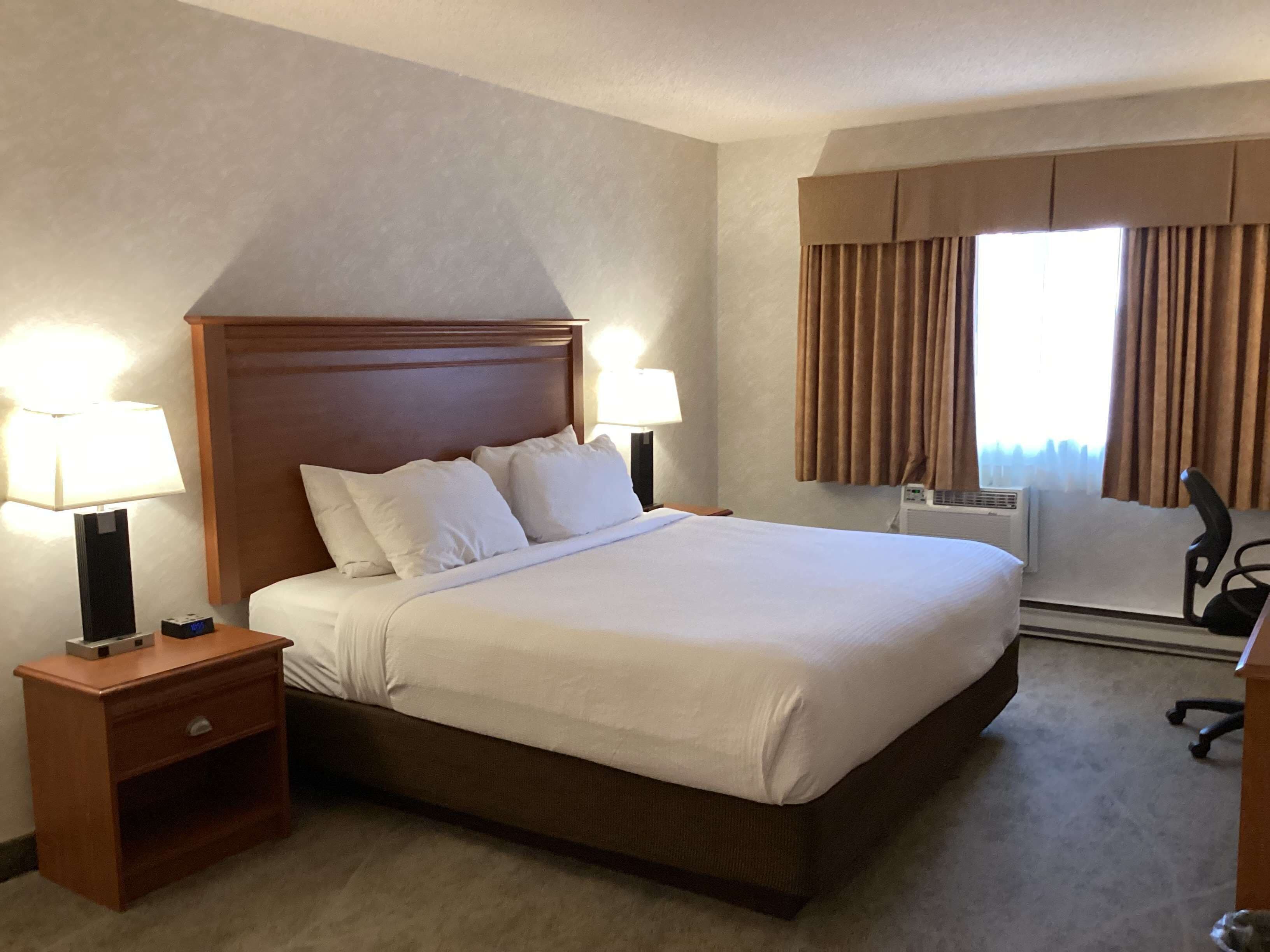 A hotel room with a large bed, two bedside lamps, a nightstand, a chair, and a window with curtains.