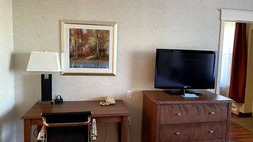 A hotel room with a desk, a chair, a television, a telephone, a painting, and a wooden dresser.
