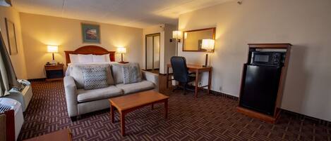 A hotel room with a sofa, a desk, a chair, a coffee table, a microwave, and a heater.
