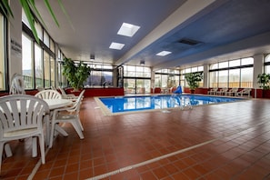 An indoor swimming pool with white chairs and tables, large windows, and potted plants.