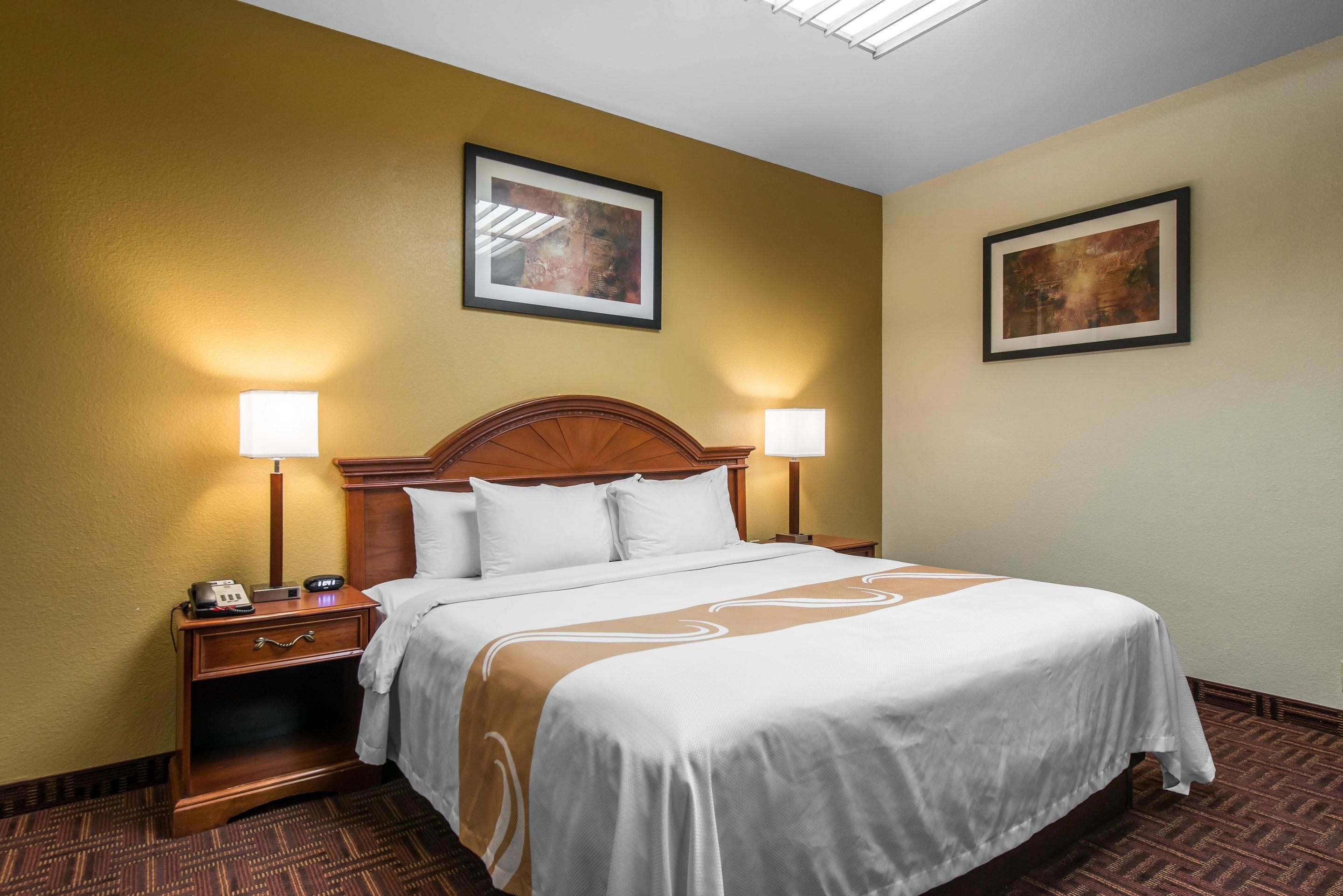 A hotel room with a large bed, two bedside tables with lamps, a wooden headboard, and two framed pictures on the walls.