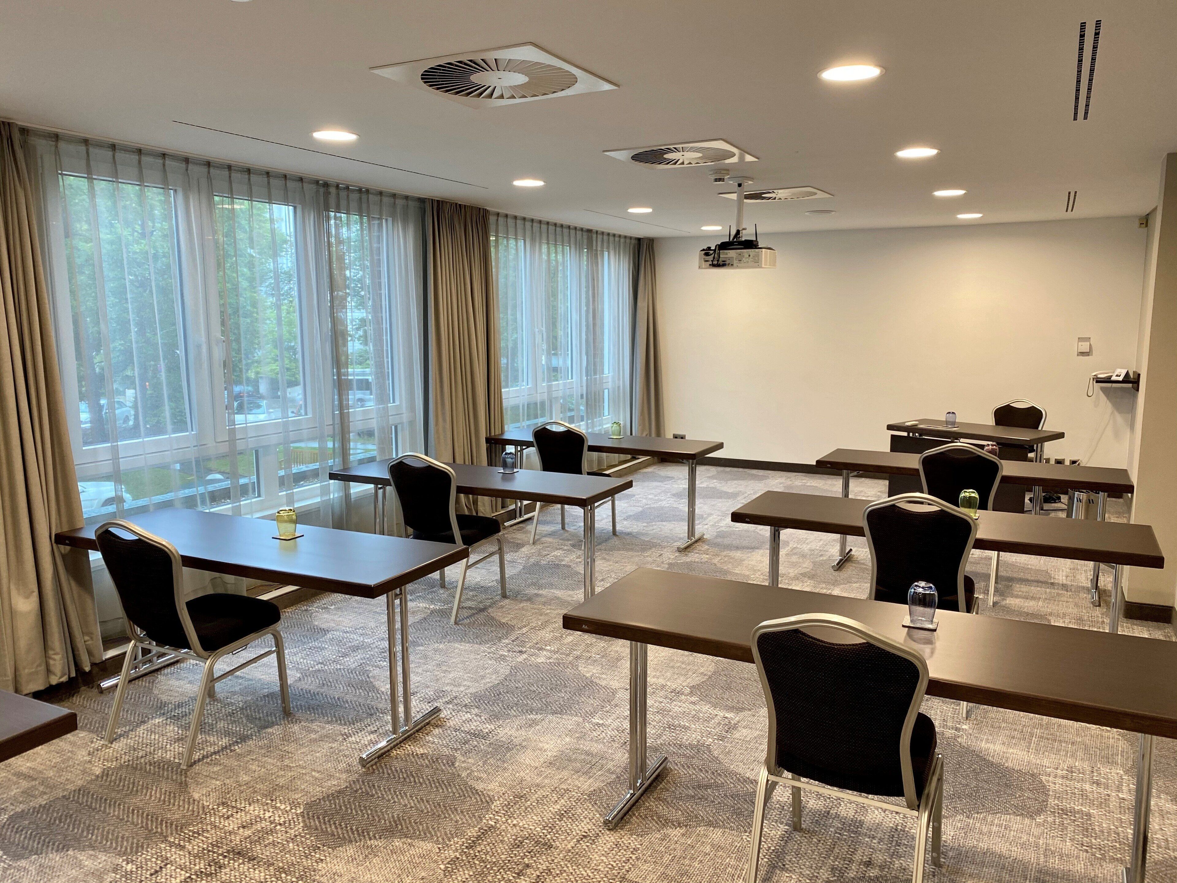 Ein Konferenzraum mit Tischen und Stühlen, großen Fenstern mit Vorhängen und Deckenleuchten.