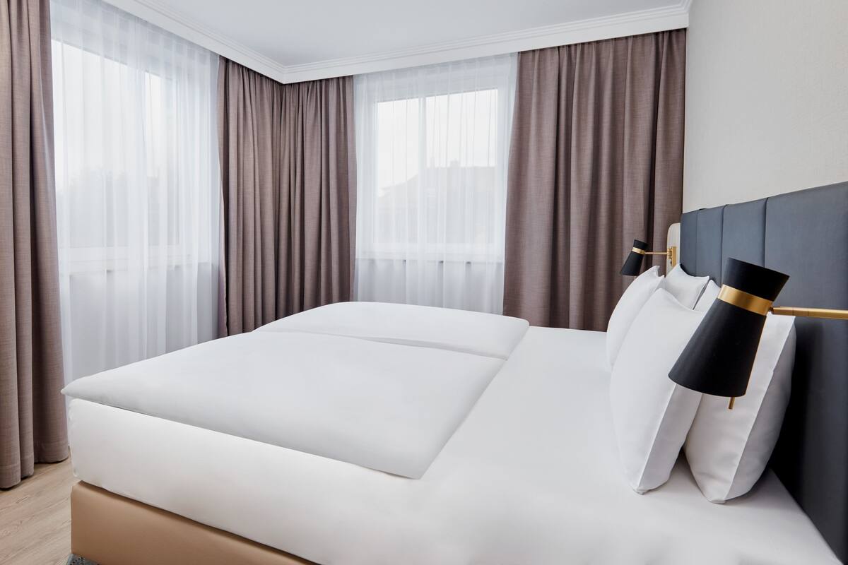 Moderni hotellihuone, jossa on suuri sänky, yöpöytä lampun kanssa ja läpikuultavat verhot.