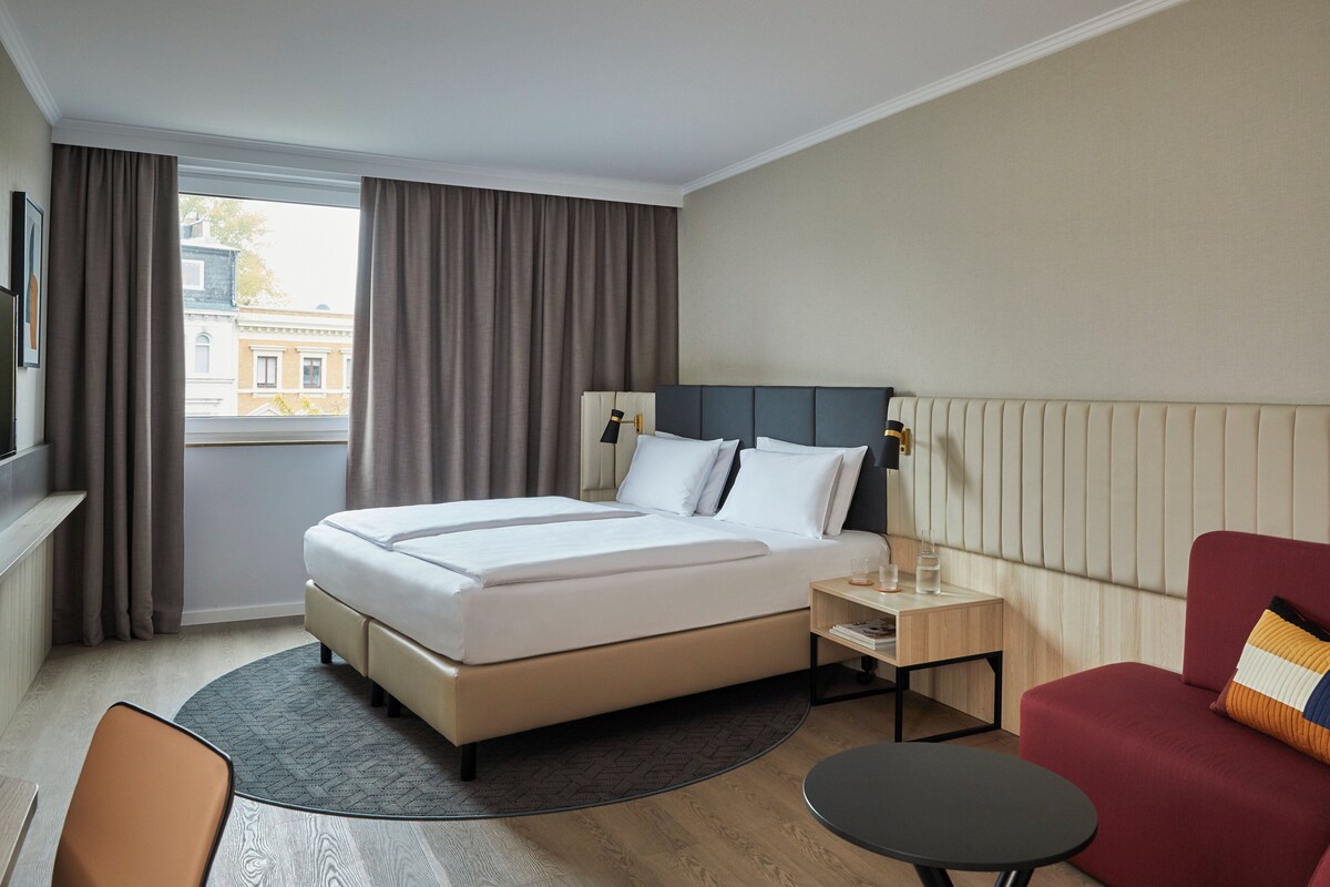 Ein Hotelzimmer mit einem großen Bett, einem Nachttisch mit Lampe, einem roten Sessel und Blick auf Gebäude.