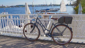 Un vélo est garé au bord de l’eau, près d’une rambarde blanche. On aperçoit l’étendue d’eau et, au loin, des voiliers.