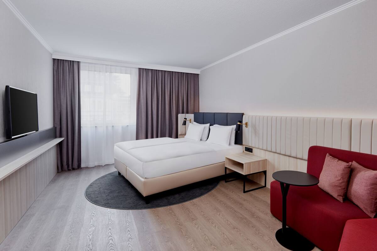 Hotellihuone, jossa on sänky, televisio, punainen sohva ja pieni pöytä.