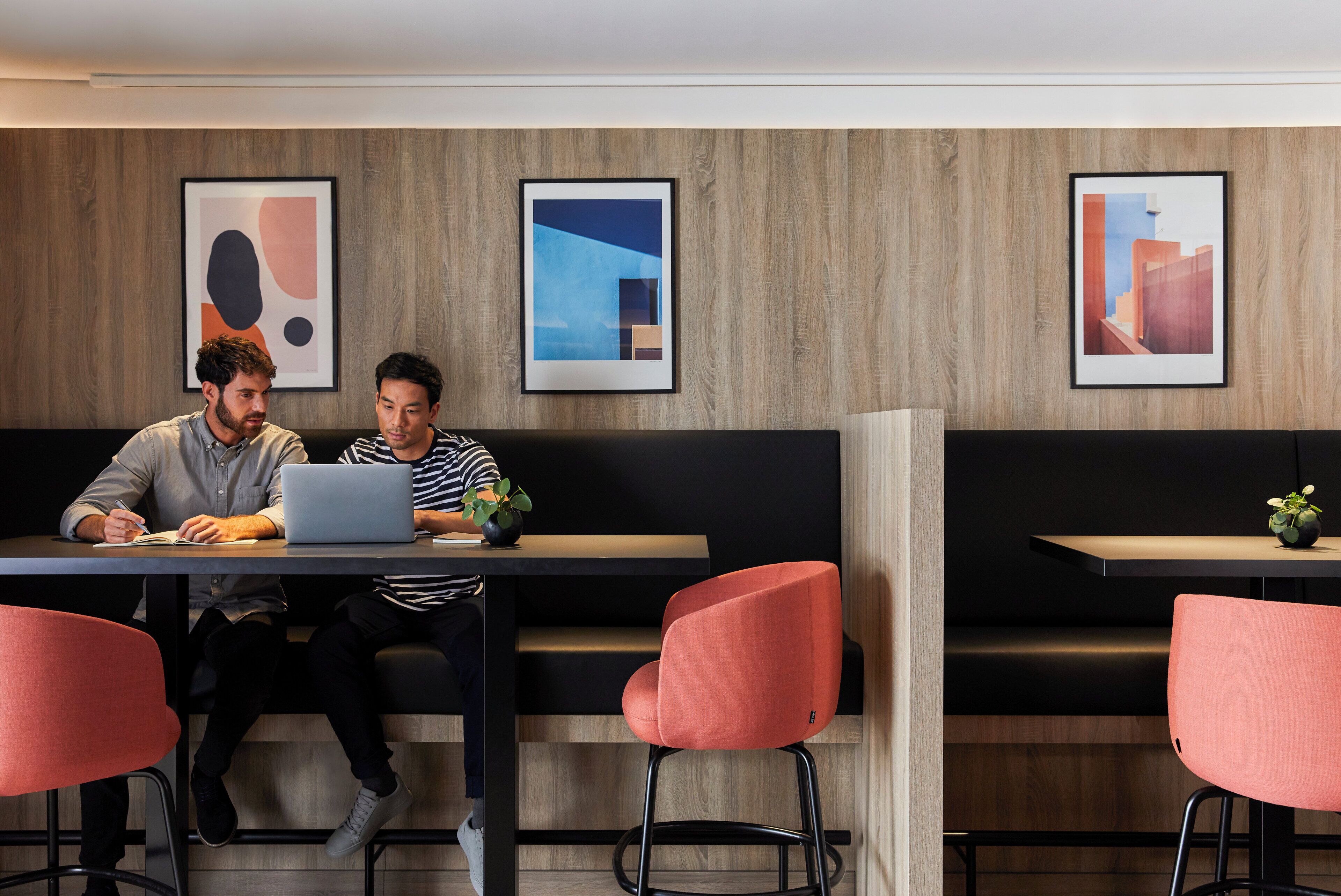 A modern interior with two individuals seated at a table with laptops, surrounded by artwork on the walls.