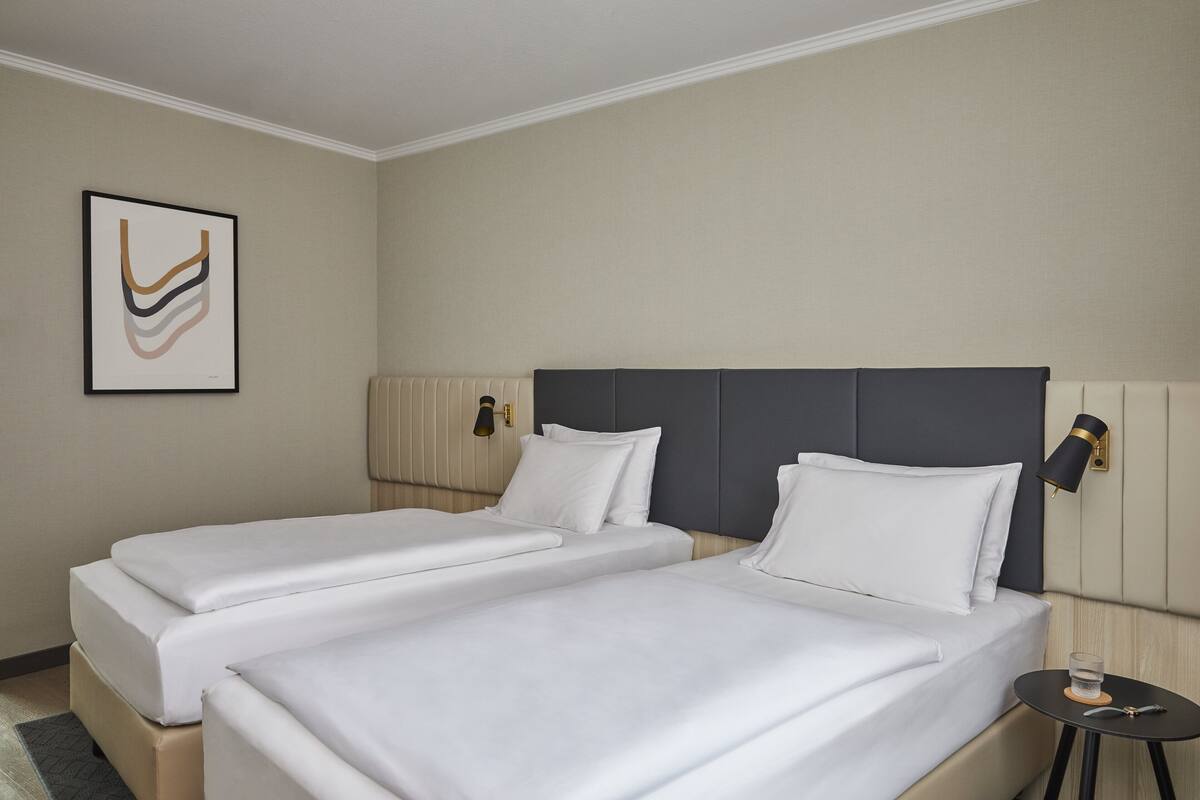 Hotellihuone, jossa on kaksi sänkyä, yöpöytä, jossa on lasi vettä, ja seinään kiinnitetty valaisin.