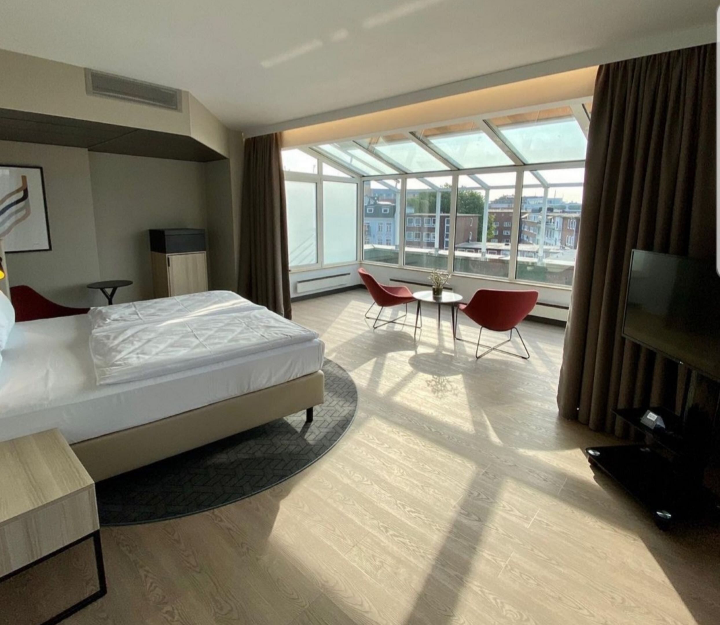 A hotel room with a bed, a TV, a desk, a chair, and a window with a view of buildings.