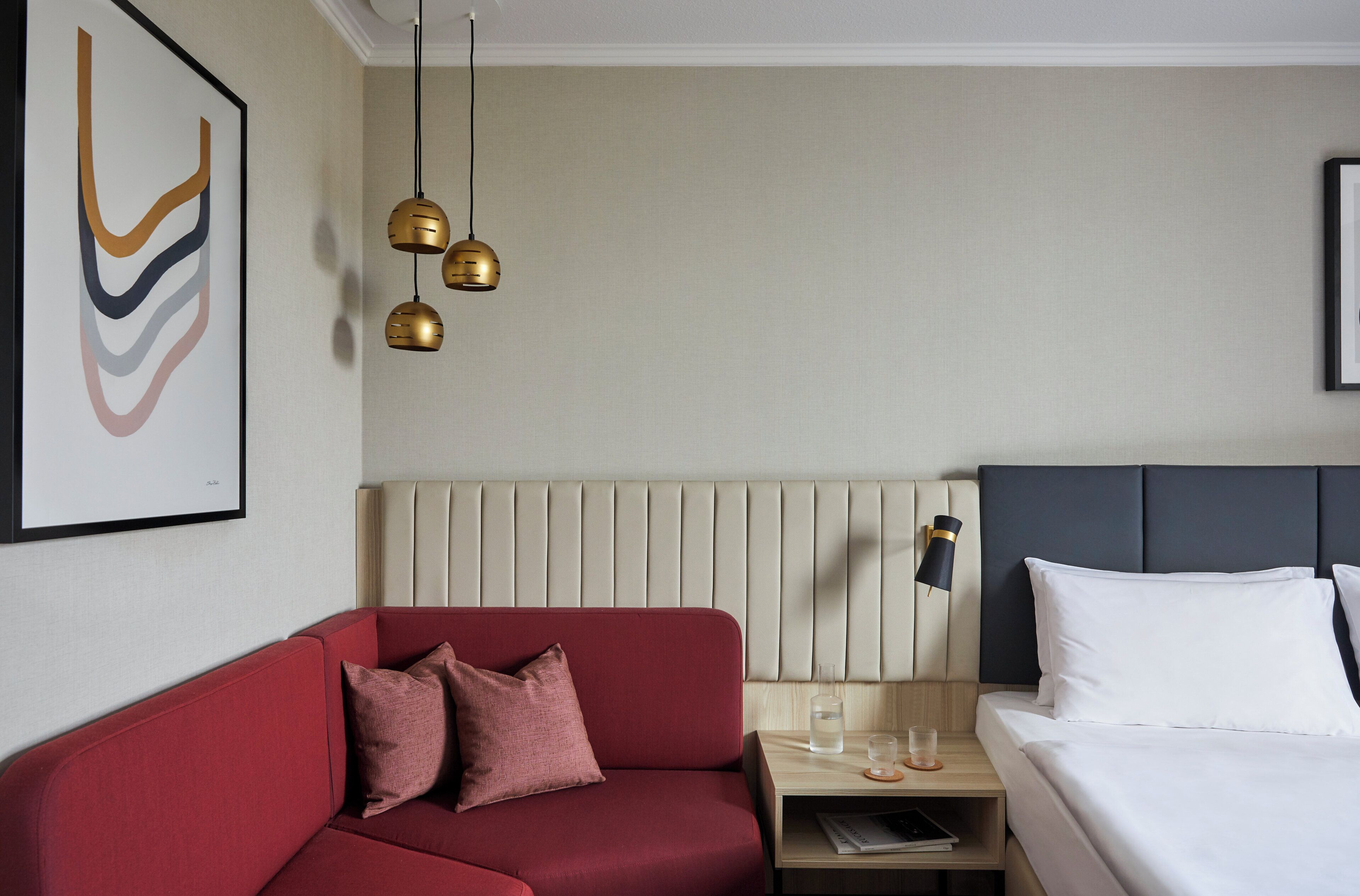 Una habitación de hotel con un sofá rojo, una cama con ropa blanca, una mesita de noche con vasos y una botella, y una lámpara fijada a la pared.