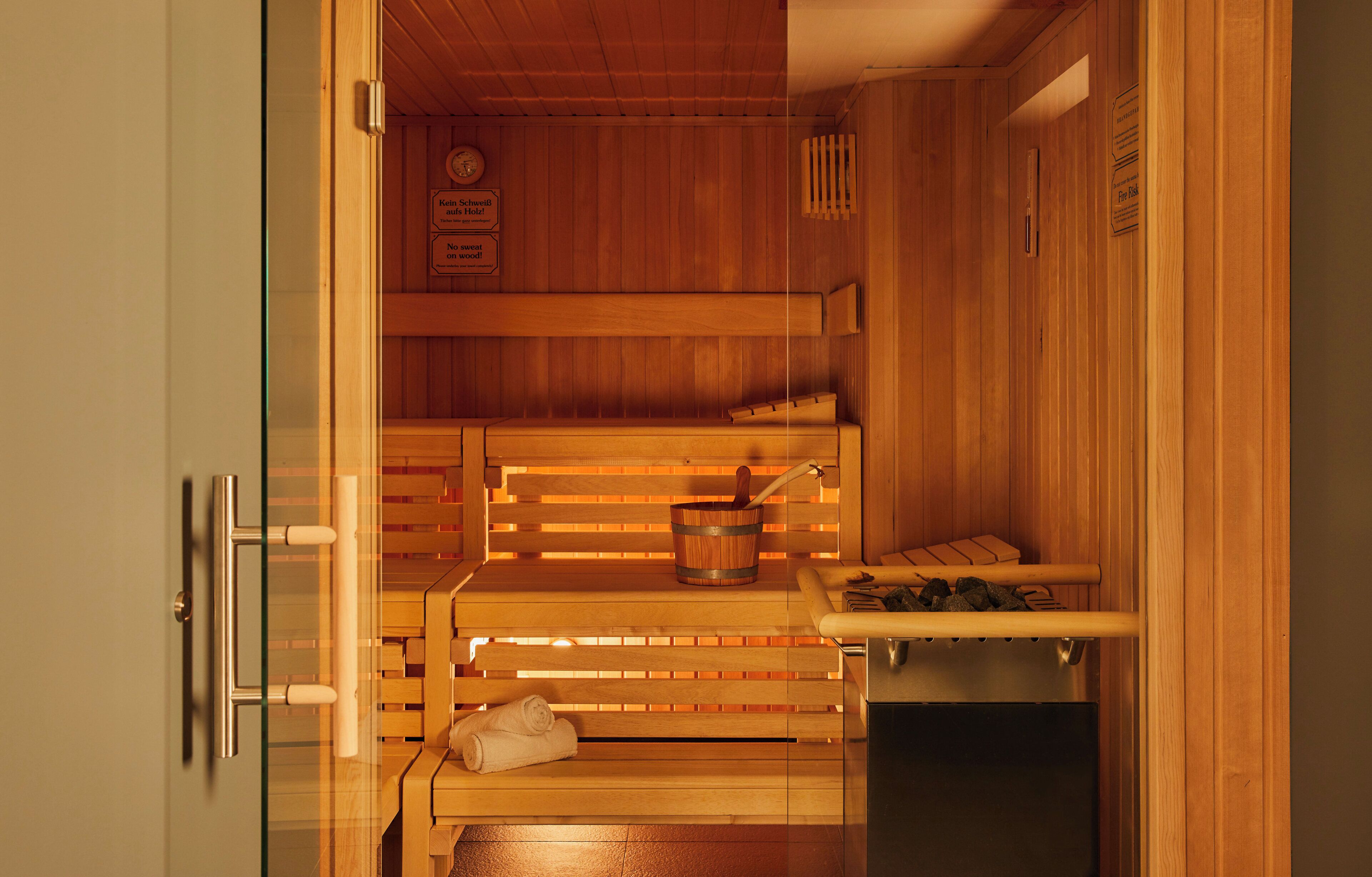 A sauna with wooden benches, a bench with a towel, a bucket with a brush, and a heater.