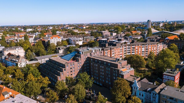Vista aérea de un paisaje urbano con una mezcla de edificios residenciales y comerciales, incluyendo un complejo de apartamentos de ladrillo rojo destacado y un edificio con techo azul.