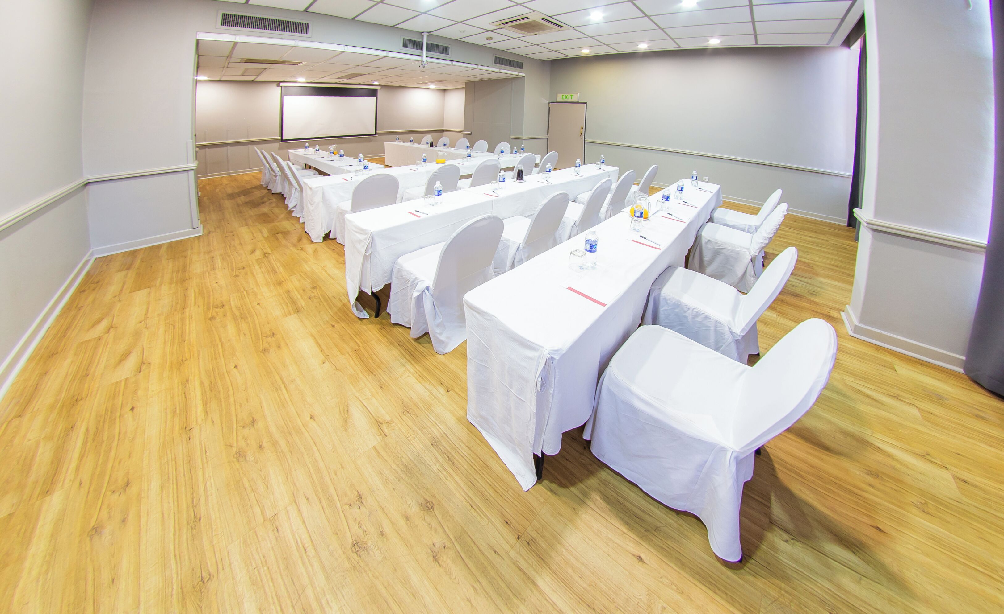 A conference room with multiple tables and chairs, a projector screen, and an exit sign.
