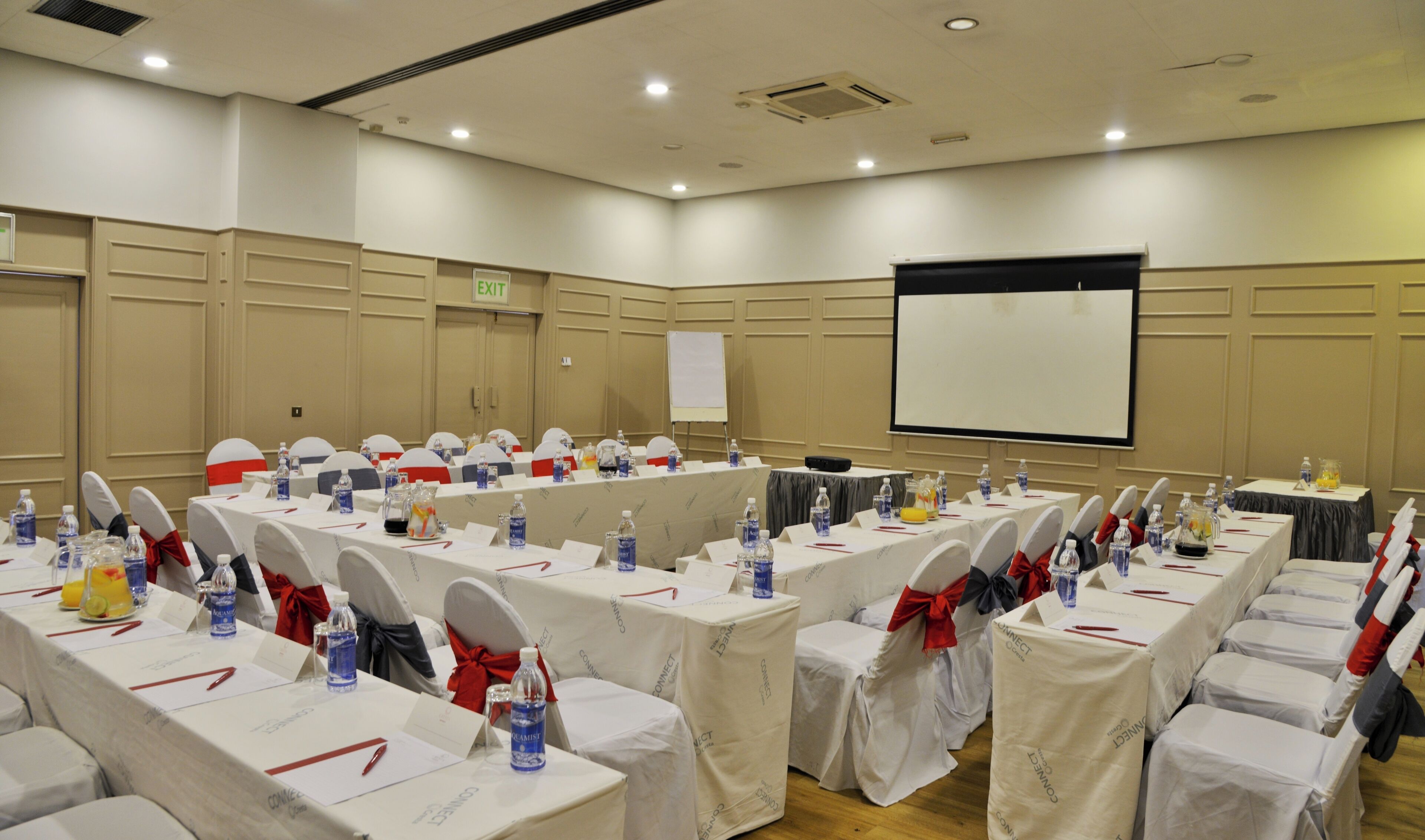 Un salón de conferencias con mesas dispuestas para una reunión, equipado con tarjetas de identificación, botellas de agua y una pantalla de proyector.