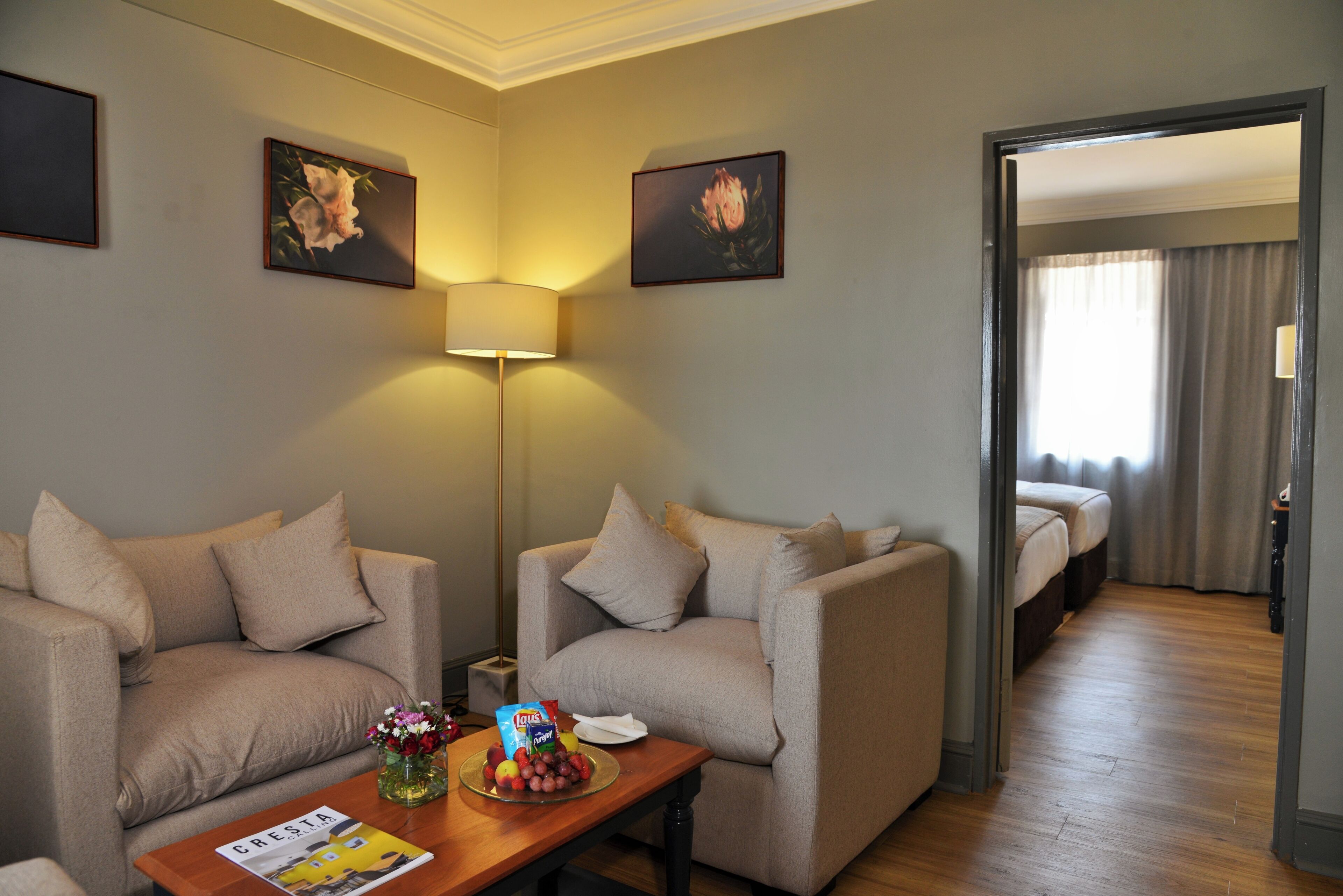 Une chambre d’hôtel avec deux canapés, une table avec un magazine et des en-cas, une lampe et deux tableaux au mur.