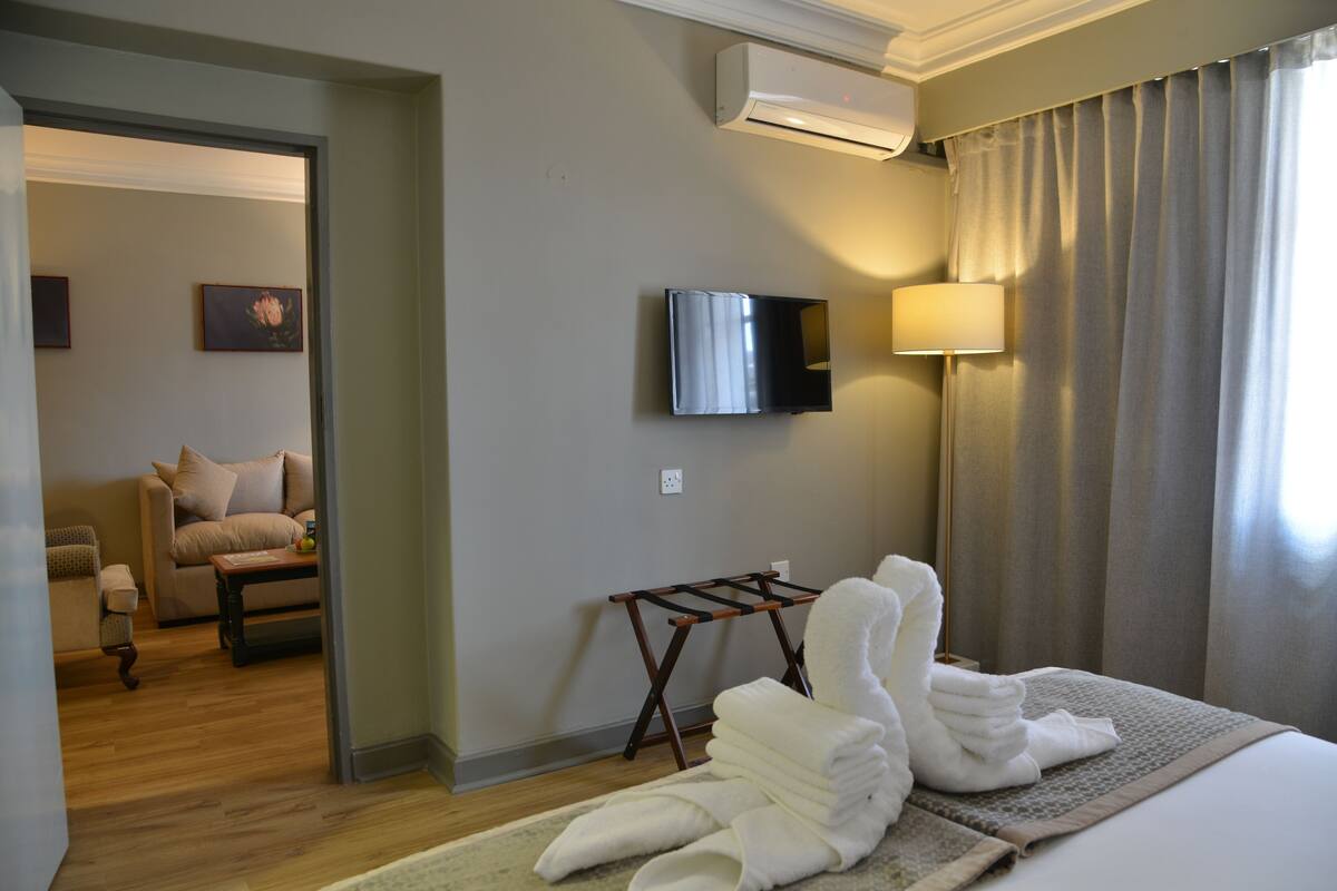 A hotel room with a bed, a TV mounted on the wall, a lamp, and a view of another room with a sofa and a painting.