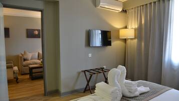 Habitación de hotel con cama, televisor montado en la pared, lámpara y vista a otra habitación con sofá y cuadro.